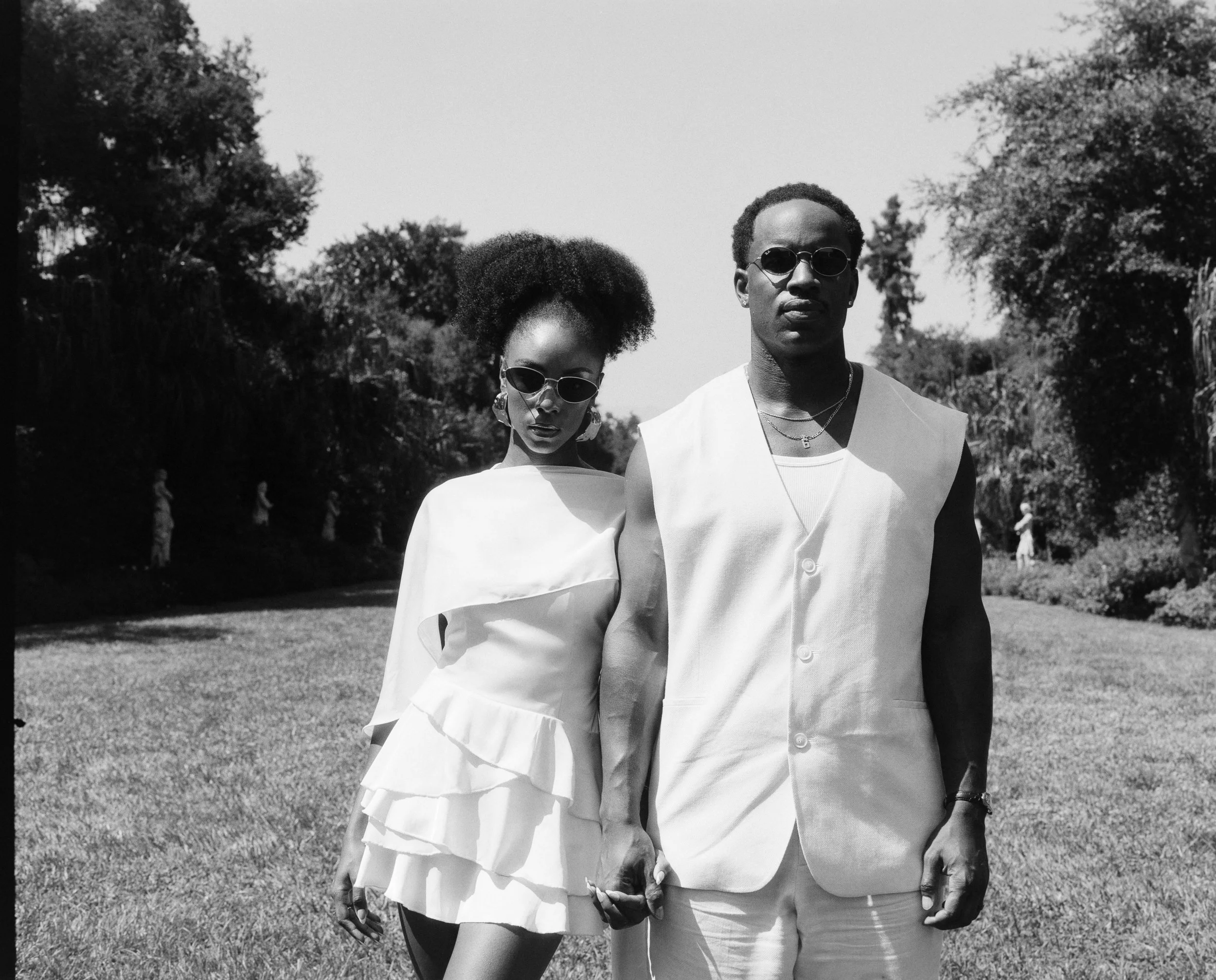 A black and white photo of a man and woman holding hands in a park. The woman has natural hair, is wearing sunglasses and a layered dress. The man has short hair, is wearing sunglasses, a sleeveless vest and pants, and has jewelry. They are standing