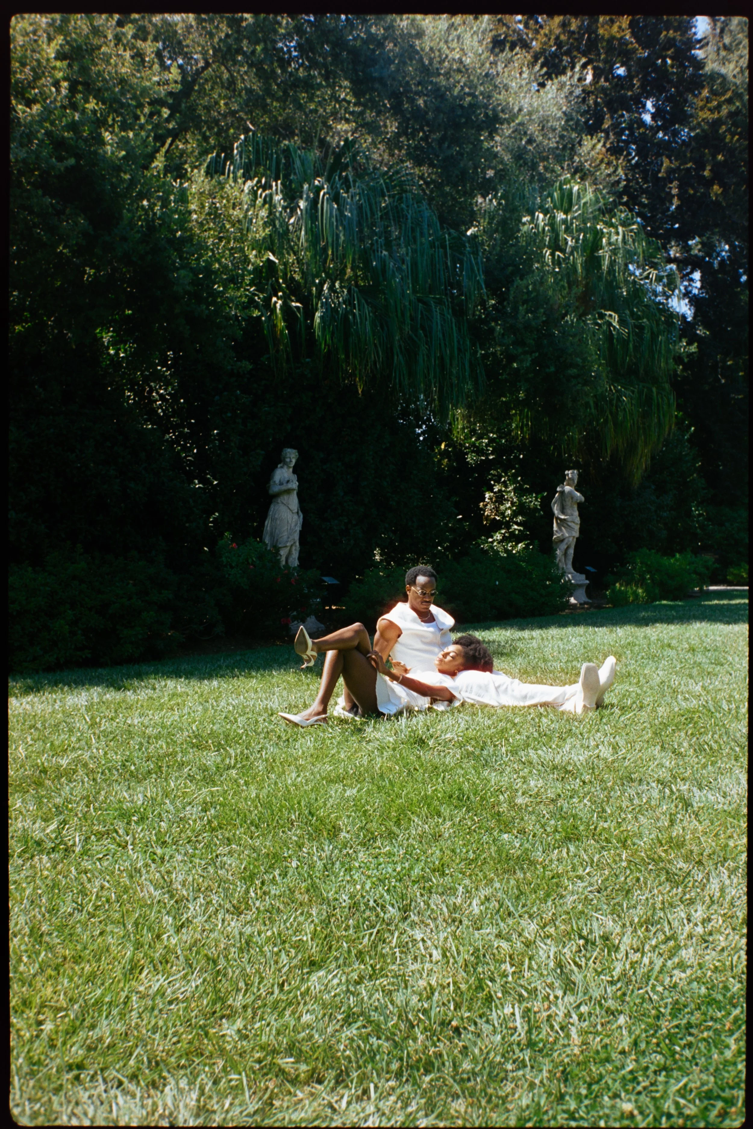 A woman and a child laying on the grass in a park with lush greenery and statues in the background.