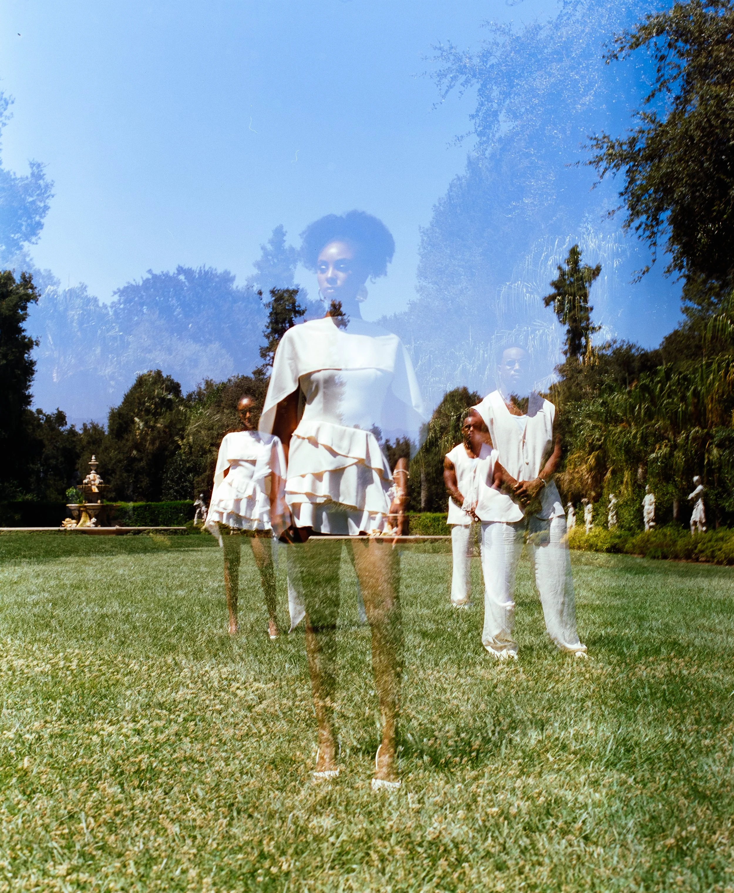A double exposure photo of women in white dresses standing on grass with trees in the background, creating overlapping images of each woman.