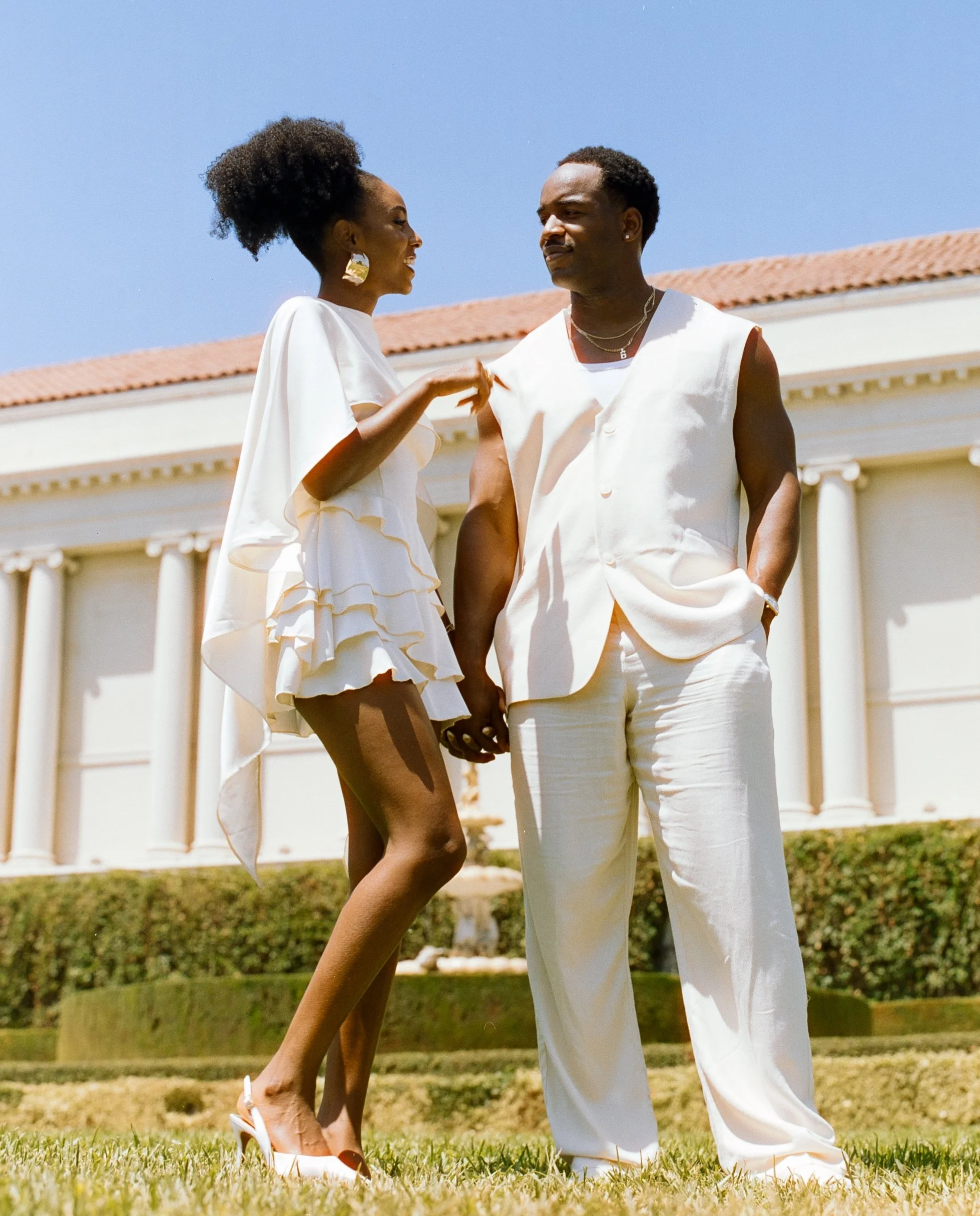 A couple holding hands and smiling at each other outside a white building with columns, dressed in white clothing, on a sunny day.