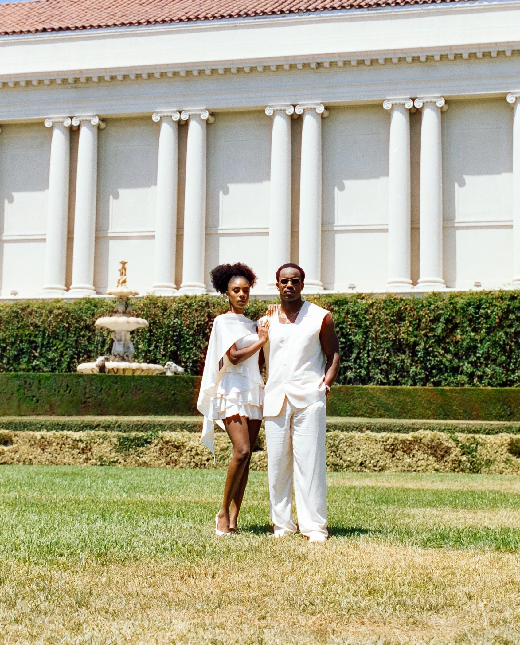 A man and woman dressed in white standing on a lawn in front of a large white building with columns and a fountain.