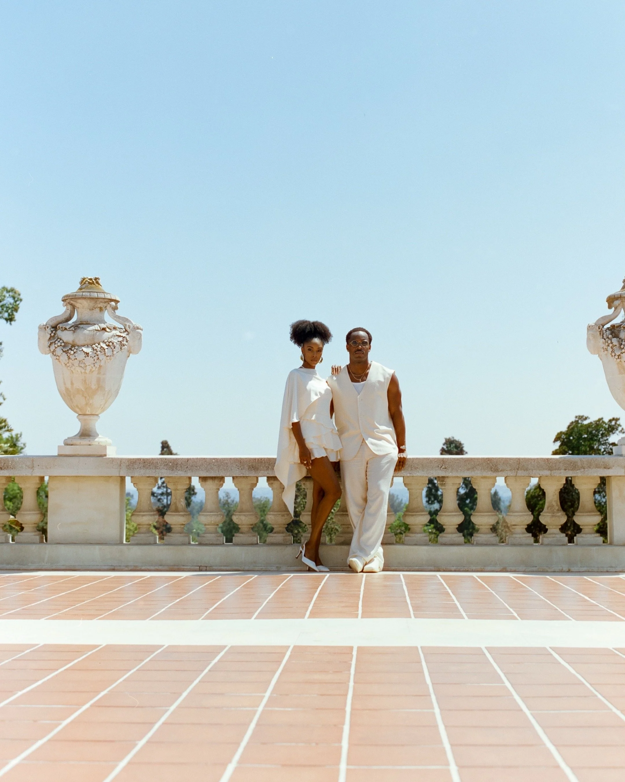 A man and woman dressed in white standing on a terrace with ornate urns and a balustrade, clear sky in the background.