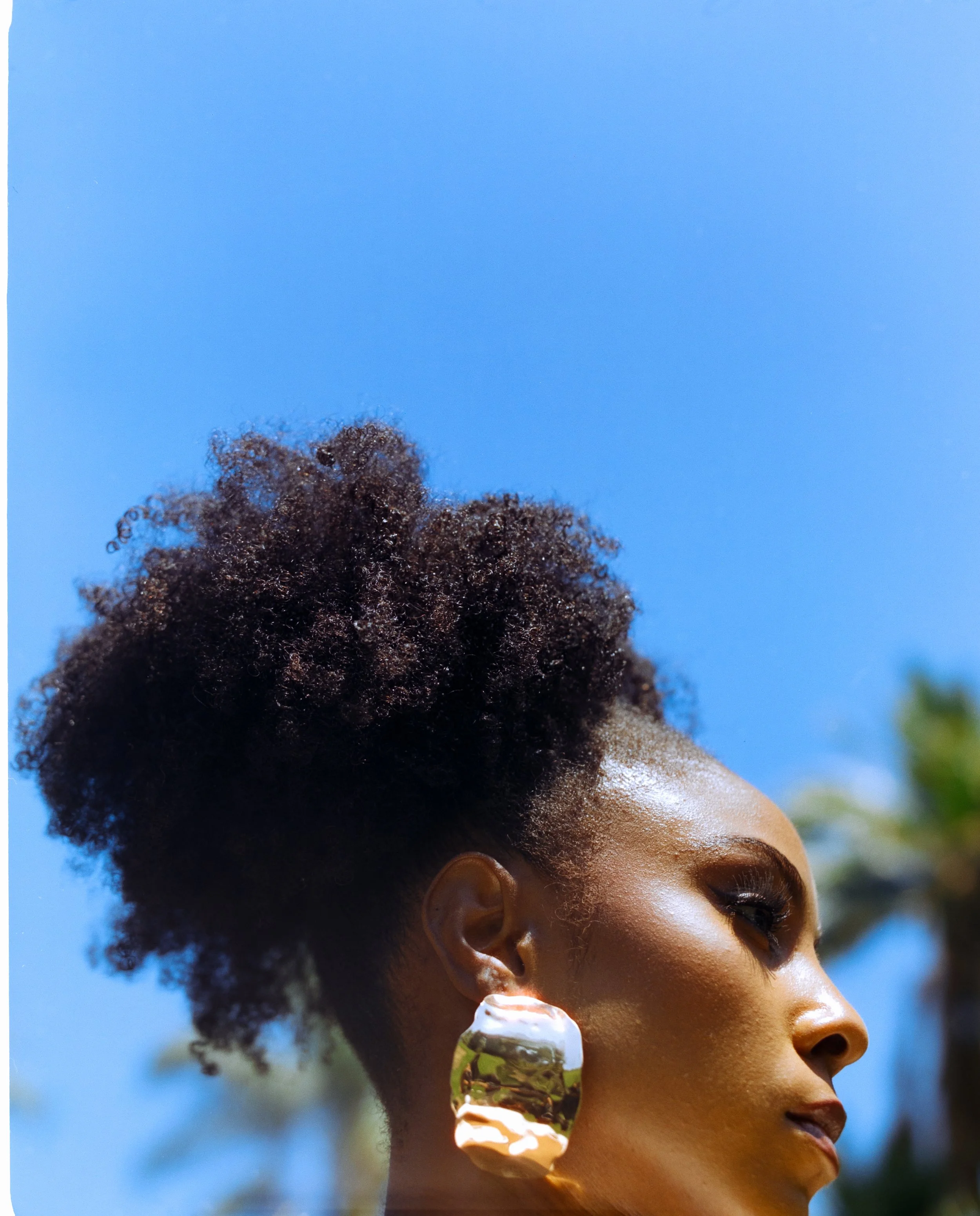 Close-up of a woman with natural curly hair styled up, wearing large reflective earrings, looking to the side against a bright blue sky with some blurred green trees in the background.