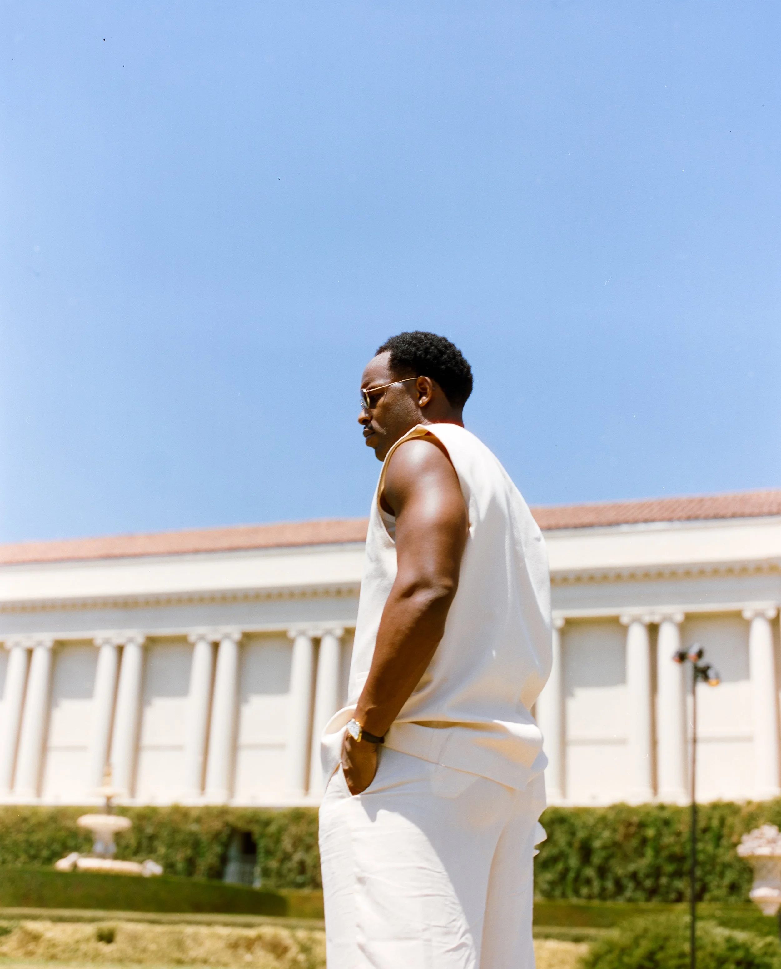 A man wearing sunglasses, a sleeveless shirt, and white pants standing outdoors with a large white building and a blue sky in the background.