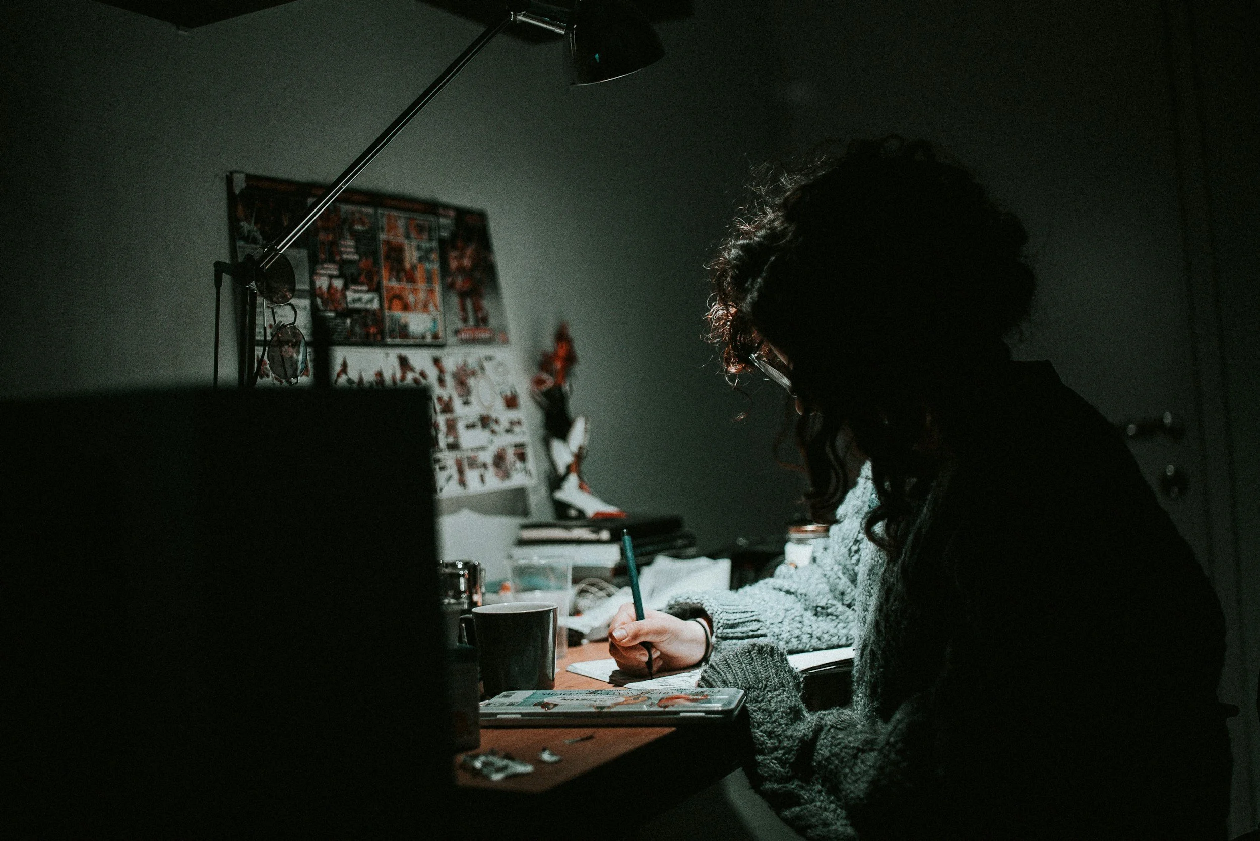 Person with curly hair writing at a cluttered desk in a dimly lit room, with a lamp overhead, a cup, and photos on the wall.