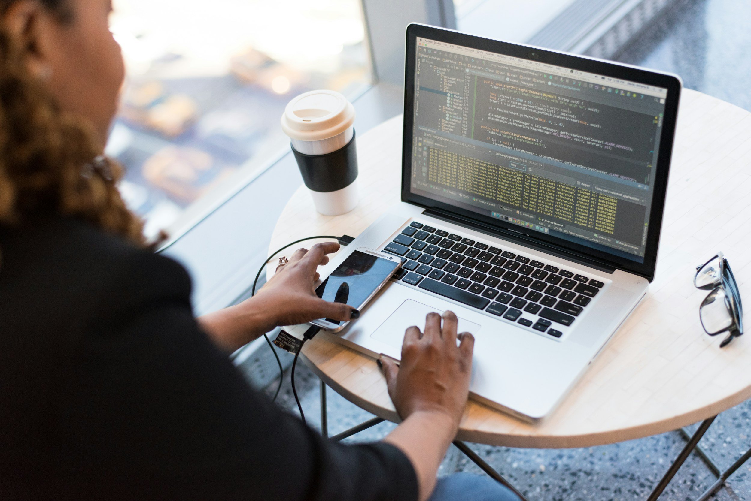 A woman working at a wooden table with a laptop displaying code, a smartphone, a pair of glasses, and a coffee cup nearby.