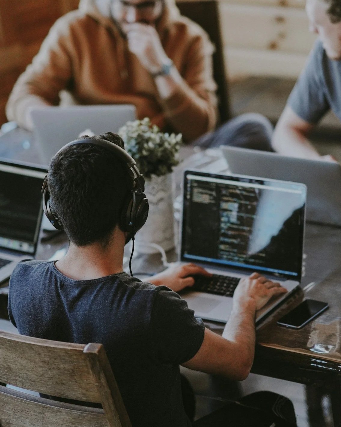 Man with headphones working on a laptop with code, at a table with smartphones and other laptops, in a casual indoor setting.
