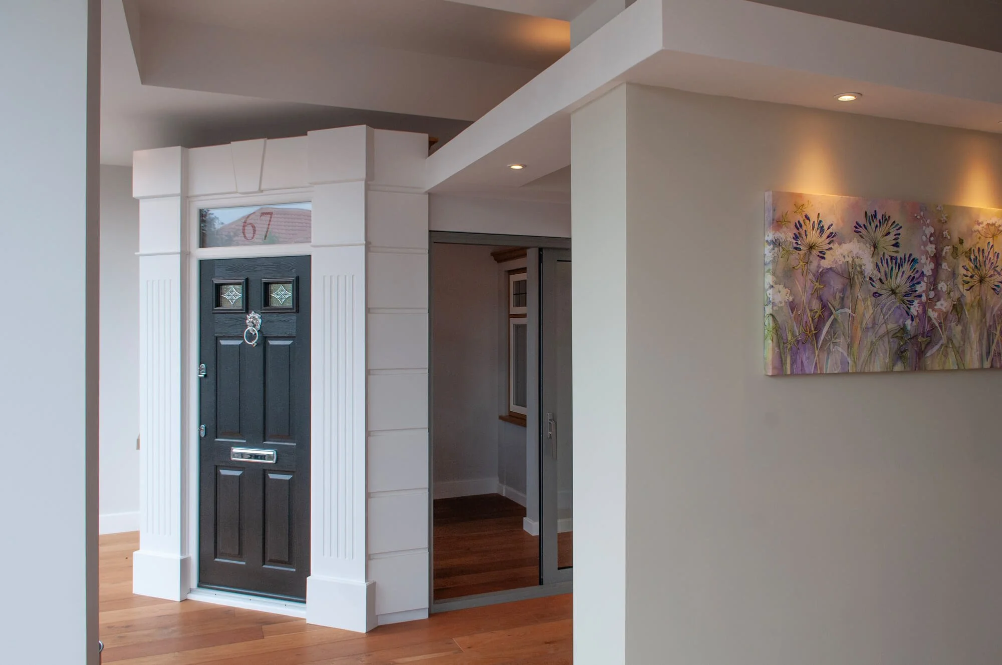 The entrance view of a home showing a black front door with decorative glass panels and a mail slot, framed by white columns and near a wall with purple floral artwork, in a room with wooden flooring.