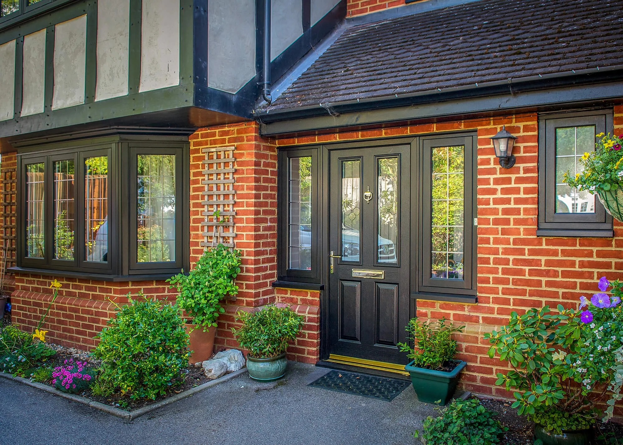 A brick house entrance with a black front door, surrounded by window frames and greenery, potted plants, flowers, and a wall-mounted lantern.