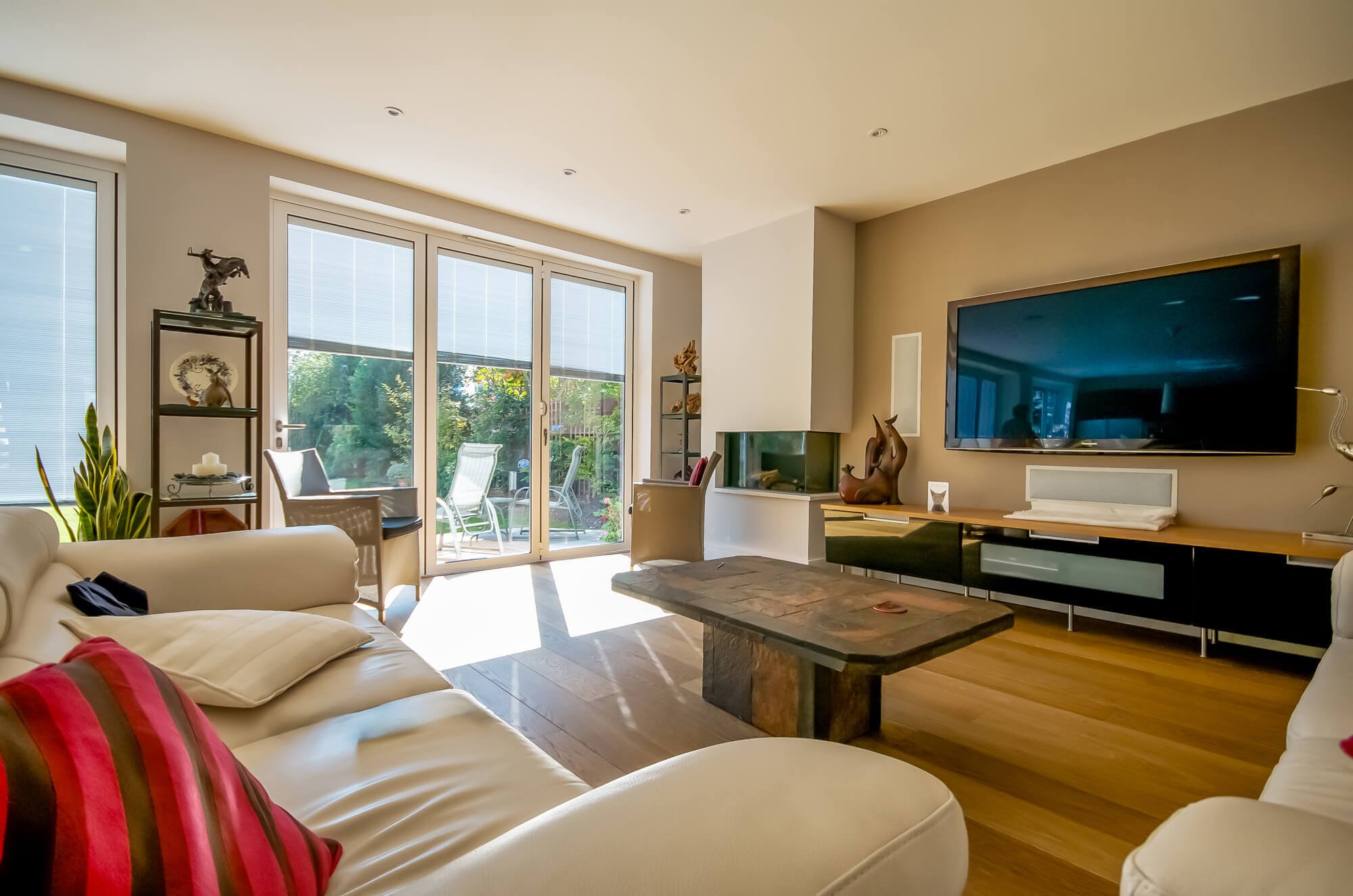Bright living room with large sliding glass doors, white couches with red and black pillows, central stone coffee table, wall-mounted TV, and patio furniture outside.