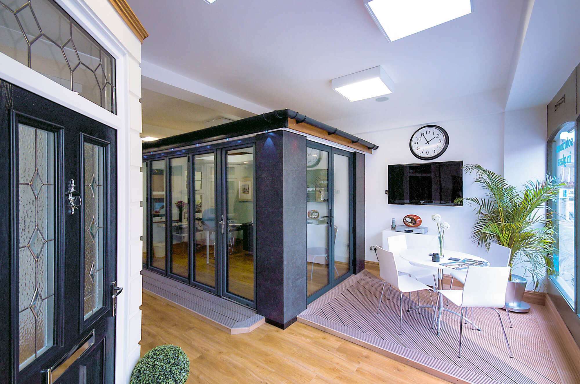 Interior view of a modern office space with a glass-enclosed meeting room, a wall-mounted TV, a round dining table with four chairs, a large potted plant, and window for natural light.