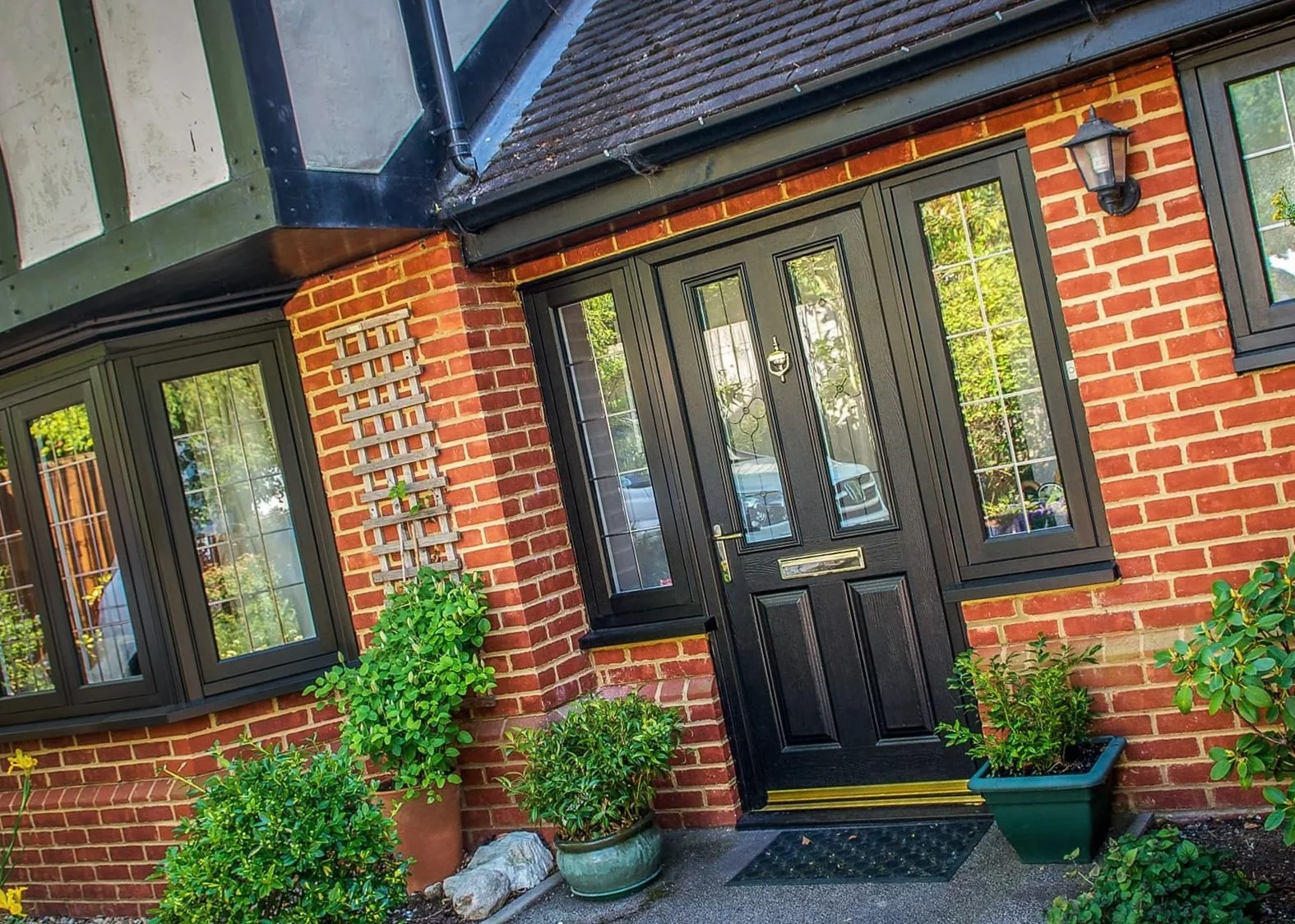 The front entrance of a brick house with a black door and black-framed windows. There are potted plants and bushes near the door, and a wall-mounted lantern light on the right side of the door.