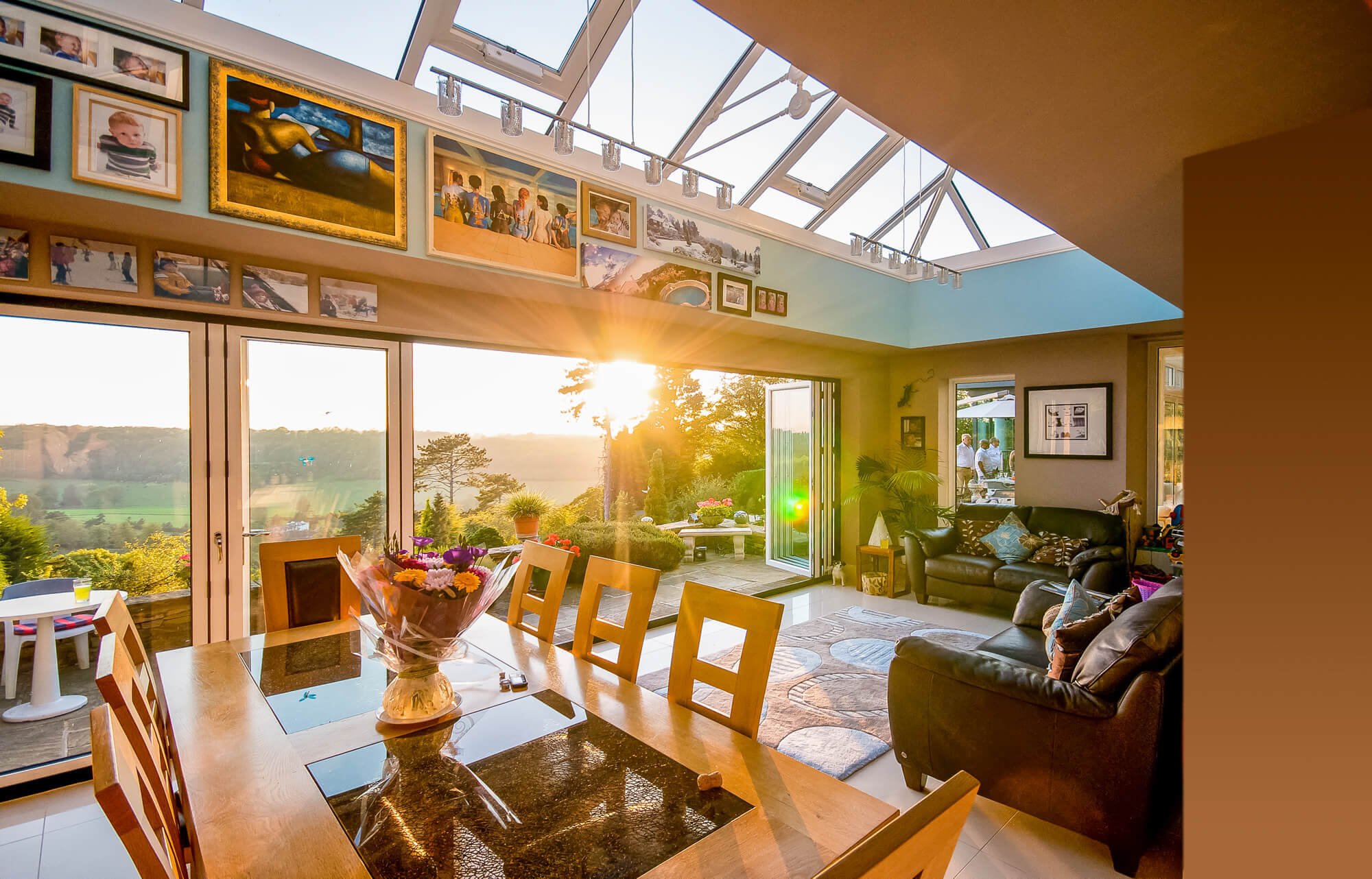 Sunset lighting inside a living room with large windows, a wooden dining table with a flower arrangement, dark leather sofas, and wall art, overlooking a lush outdoor landscape with trees.