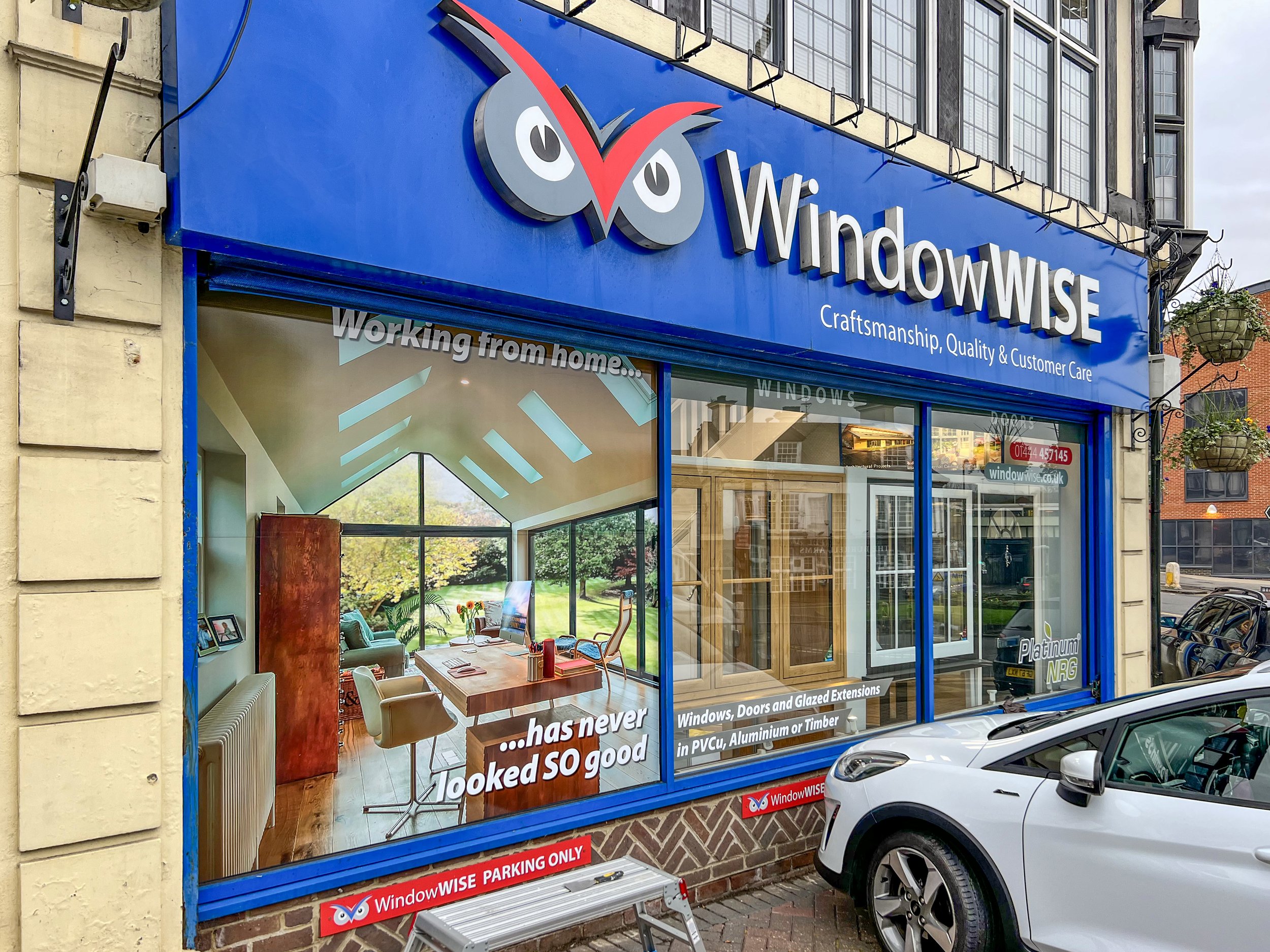  storefront with a large blue sign reading 'WindowWISE' alongside an owl logo, window and door displays, a poster inside showing an office space with large windows, and parked cars outside.