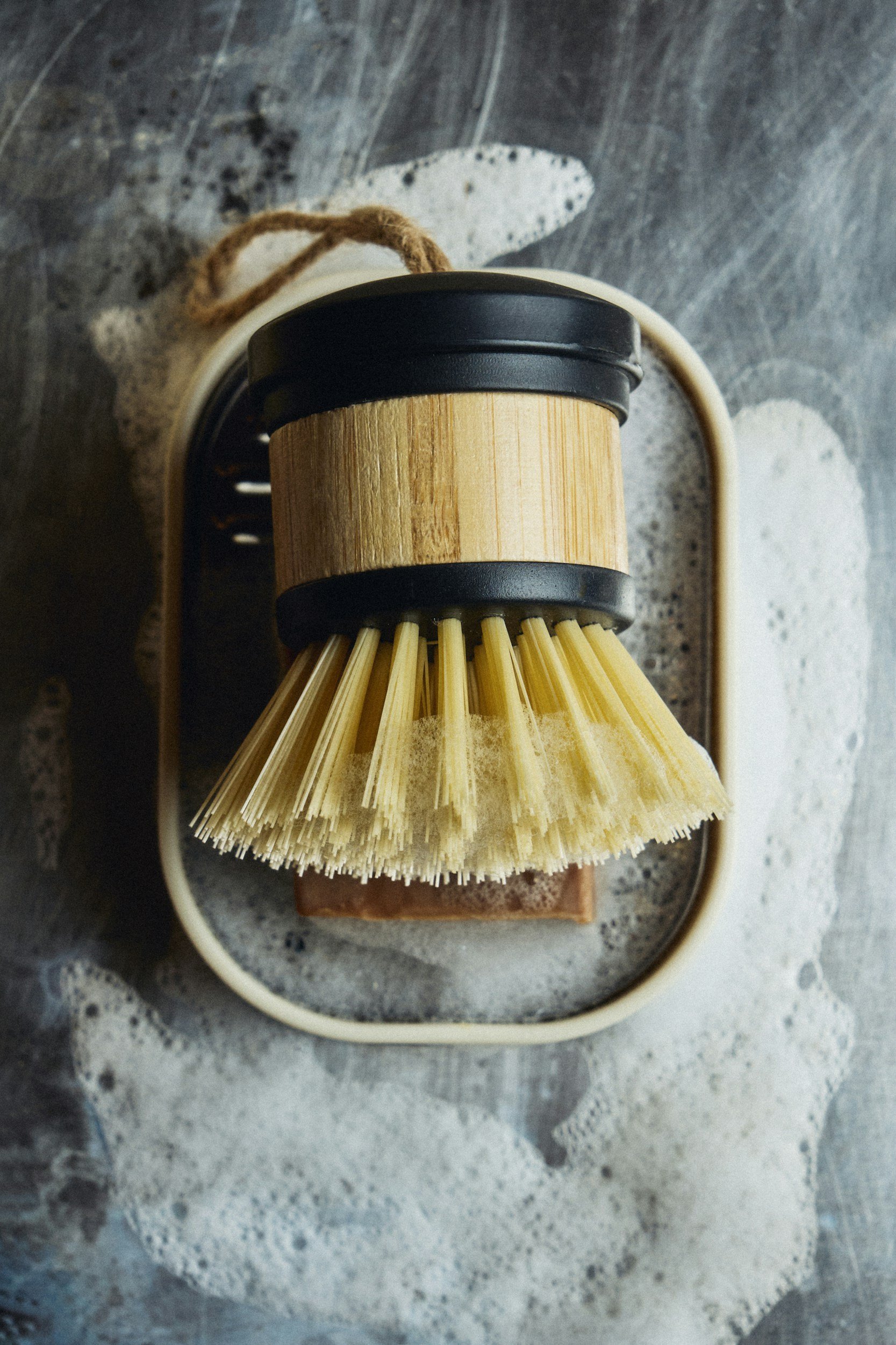 A wooden dish brush and a soap dish are arranged on a bathroom countertop with soap suds and foam.