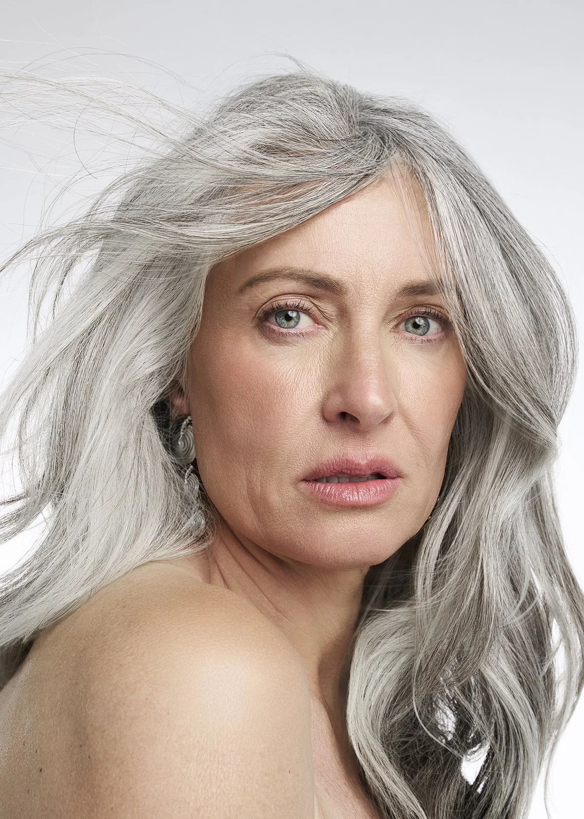 A close-up portrait of an older woman with long, wavy gray hair and light skin, looking at the camera with a neutral expression.