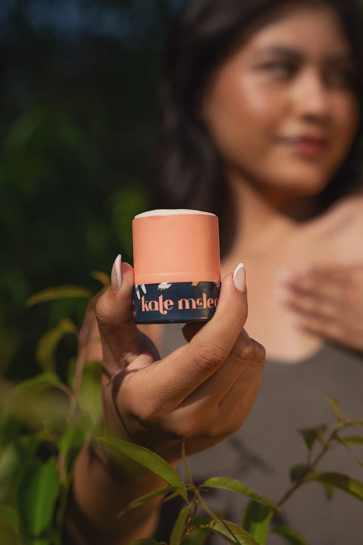 A woman holding a small, round container of skincare product labeled 'kale mlep' with greenery in the background.