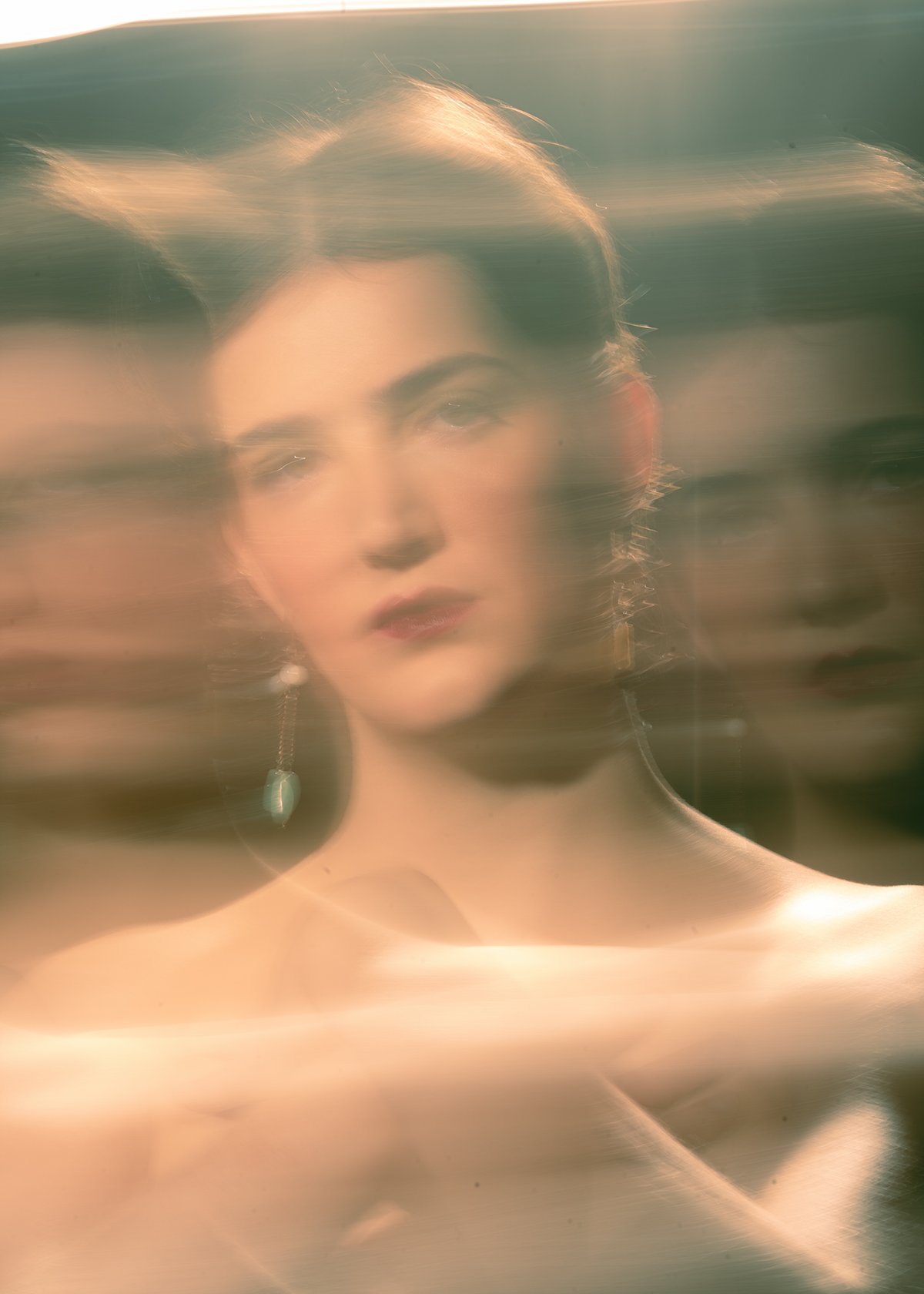 A blurry, artistic portrait of a woman with short hair, wearing earrings, with sunlight and horizontal streaks across the image.