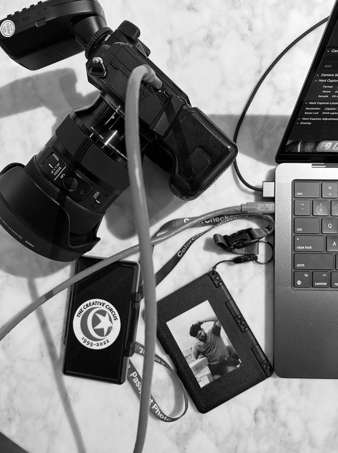 A collection of photography and tech equipment on a white surface, including a camera, a laptop, a memory card case, a photograph on a small device, and various cables.