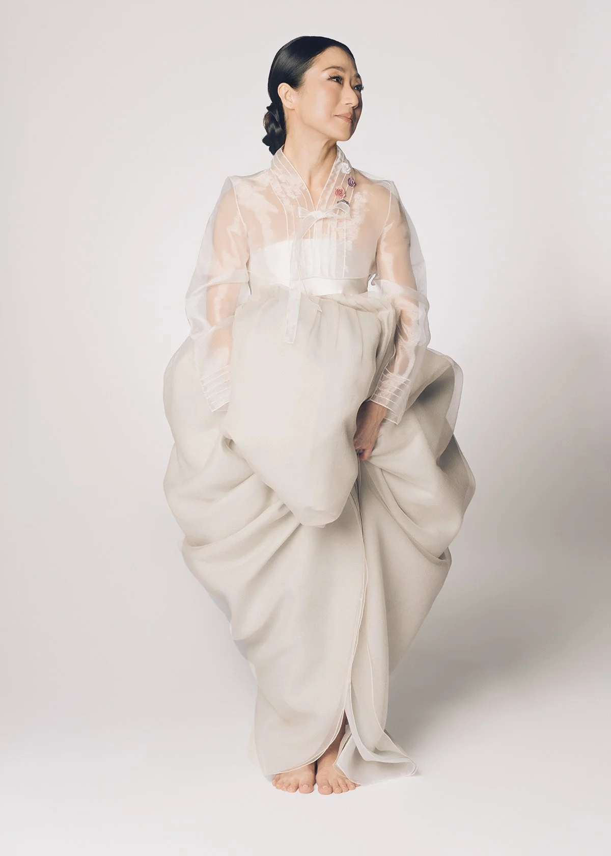 A woman with dark hair styled in an updo, wearing a sheer blouse with embroidered roses and wide, flowing cream-colored pants, standing barefoot against a plain light background.