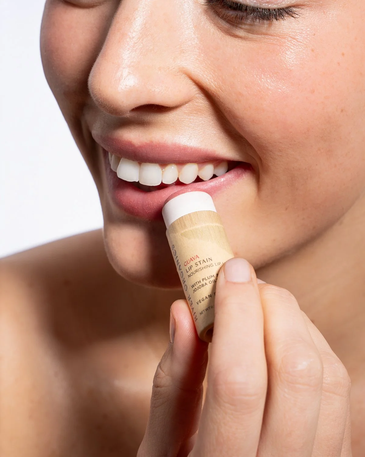 Close-up of a smiling woman applying lip balm to her lips with a stick labeled 'CUAVA' featuring natural ingredients like plum oil and jojoba oil.