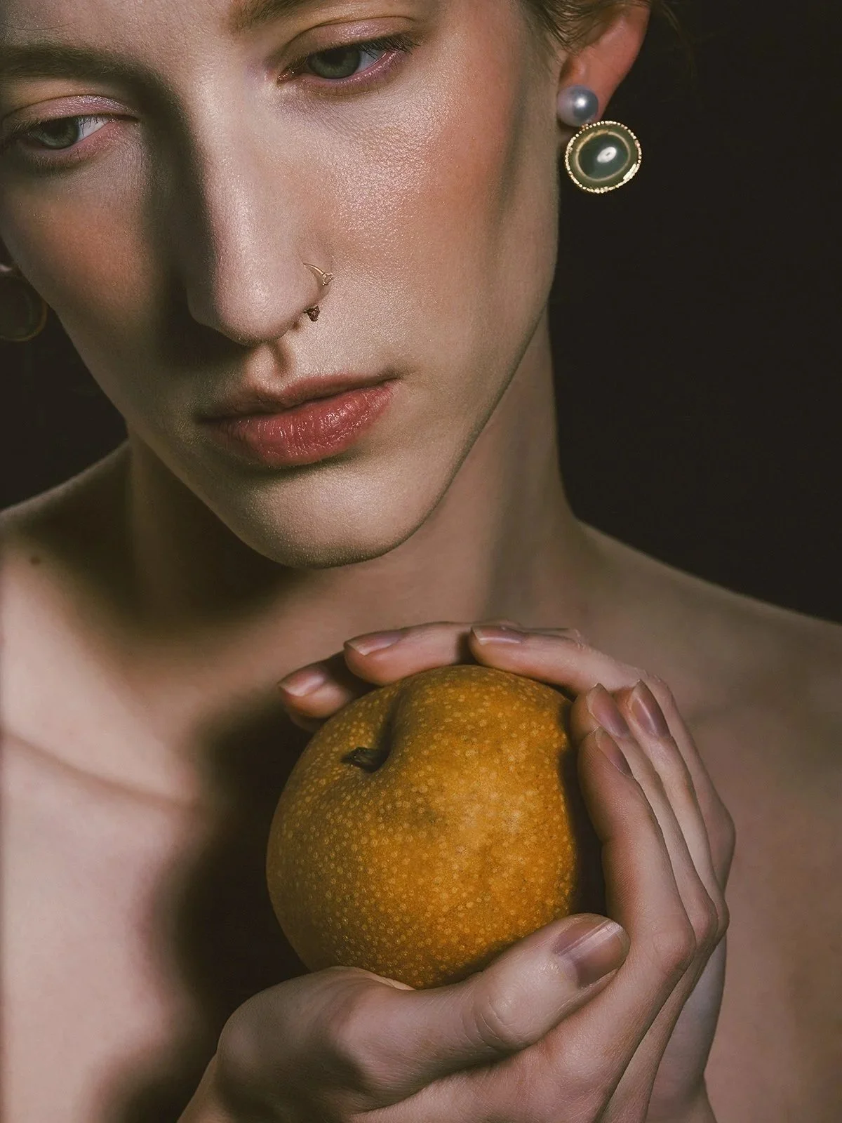 A close-up of a woman with fair skin and blue eyes holding a yellow-orange pear against her chest. She has pearl and decorative earrings, a subtle nose ring, and is looking downward with a calm expression against a dark background.