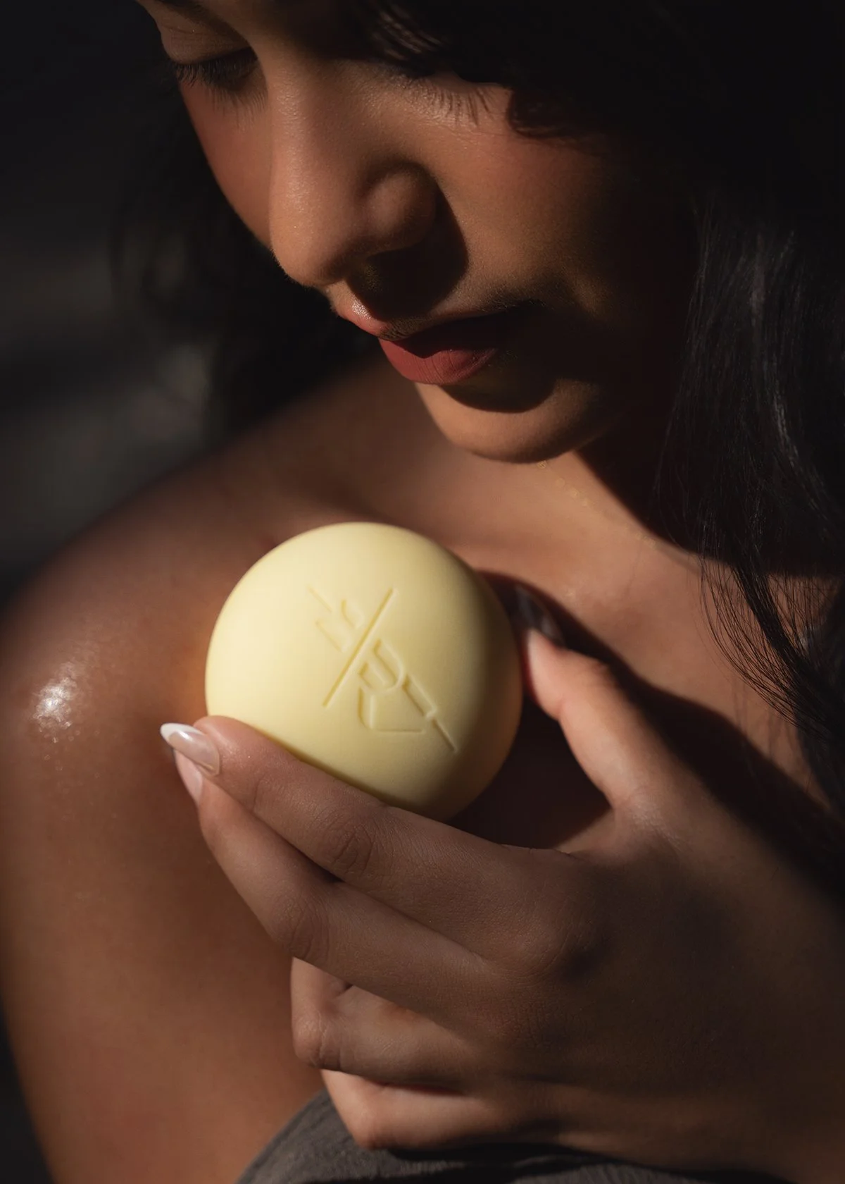 A woman with dark hair and smooth skin holding a white heart-shaped object with an engraved logo close to her shoulder. The woman is looking down at the object, and her face is partially visible, with soft lighting highlighting her features.