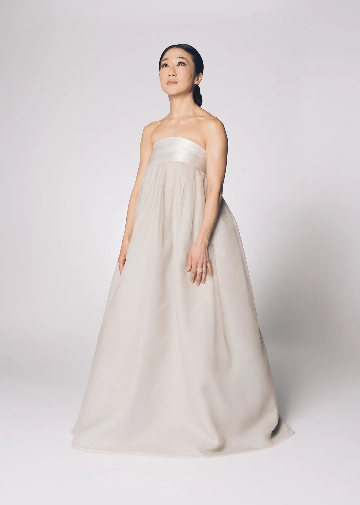 A woman with dark hair styled in a low ponytail wearing a strapless white gown with a fitted bodice and full skirt, standing against a plain light background.