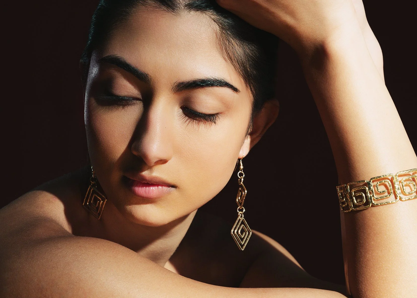 Close-up of a woman with closed eyes wearing gold earrings and a gold bracelet, with one arm raised and her hand resting on her forehead.