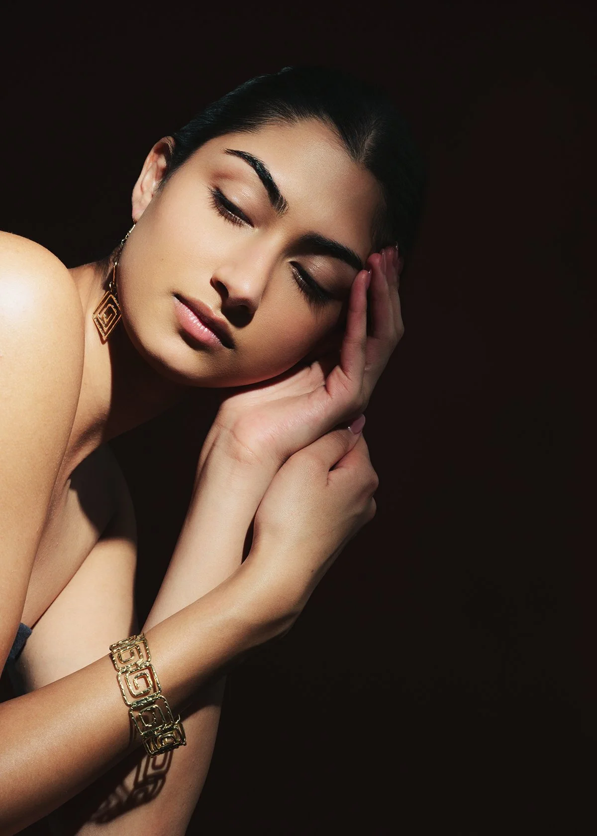 A woman with closed eyes, resting her face on her hand, wearing gold jewelry, against a dark background.