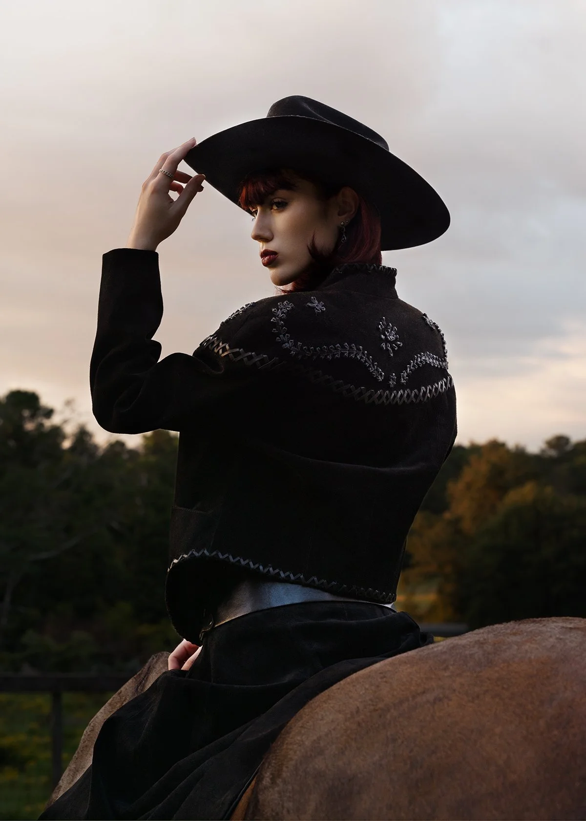 A woman wearing a wide-brimmed hat, black embroidered jacket, and dark pants, sitting on a horse outdoors during sunset.
