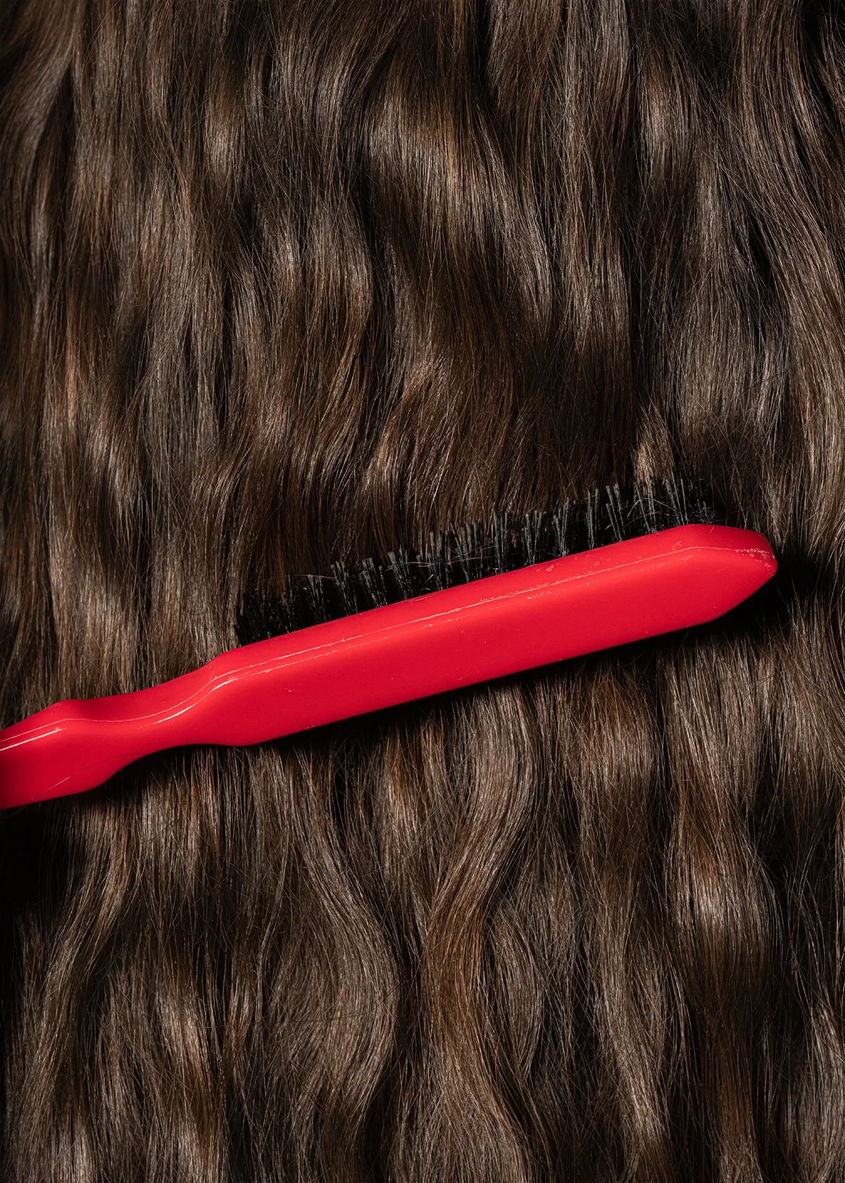 A red hairbrush combs through brown, wavy hair.