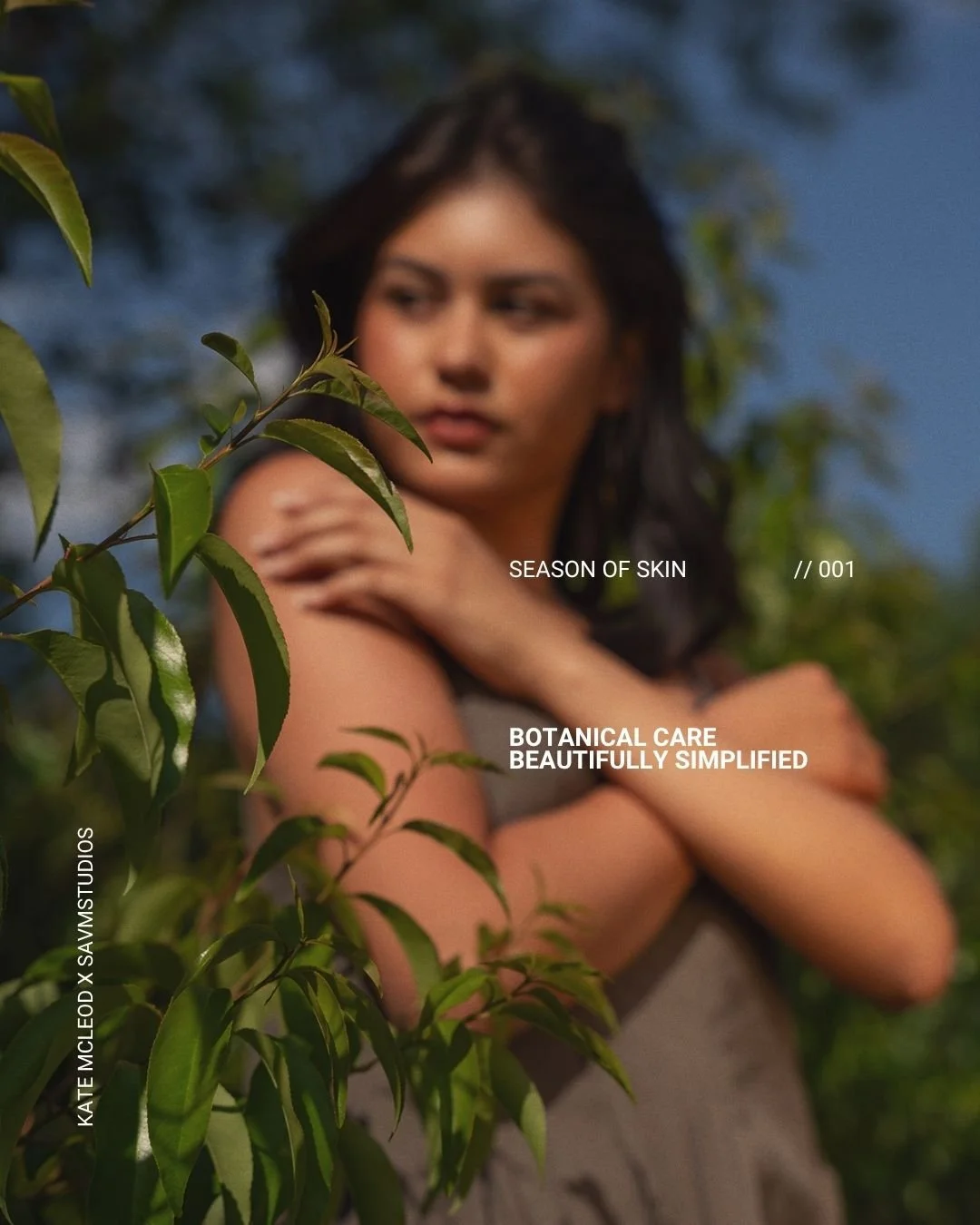 A woman with dark hair standing outdoors, partially obscured by green leaves, with a blurred background of trees and sky, promoting botanical skin care.