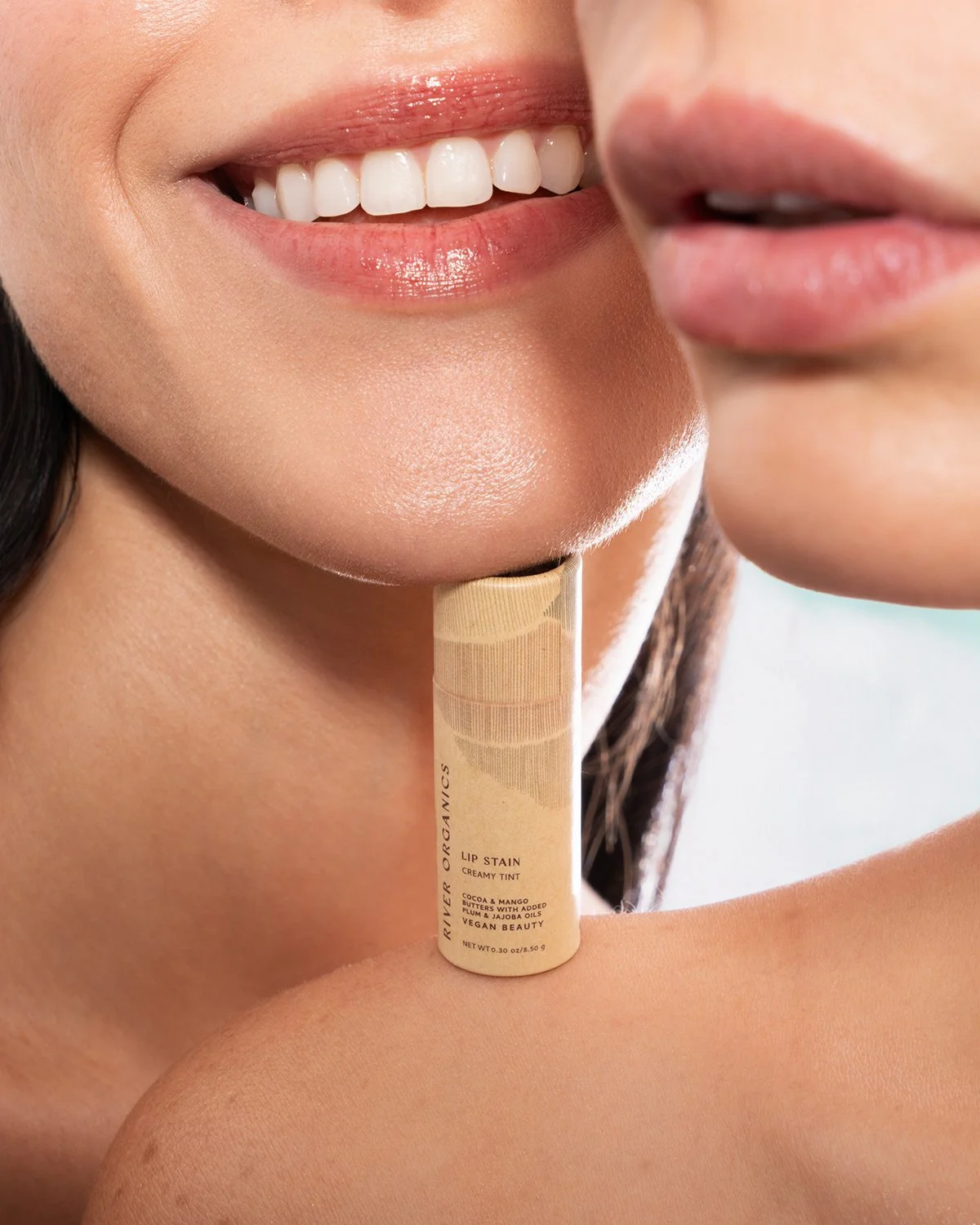 Close-up of two smiling women showing their teeth, one with glossy lips, and a lipstick tube labeled "River Organics Lip Stain Creamy Tint" positioned beneath the chin of one woman.