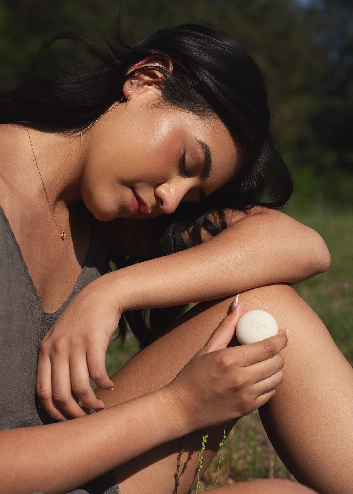 A woman with dark hair and jewelry is outdoors, holding a round soap on her shoulder with her eyes closed and a peaceful expression.