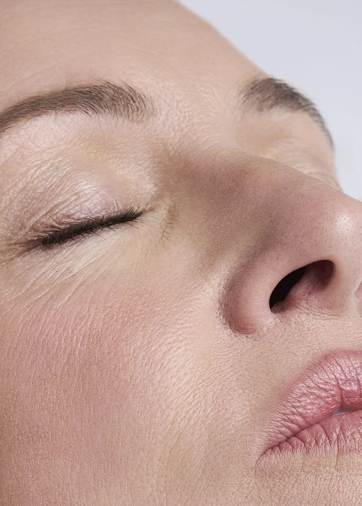 Close-up of a person's face with closed eyes, showing smooth skin, well-groomed eyebrows, and natural lip color, focusing on the nose and cheek area.