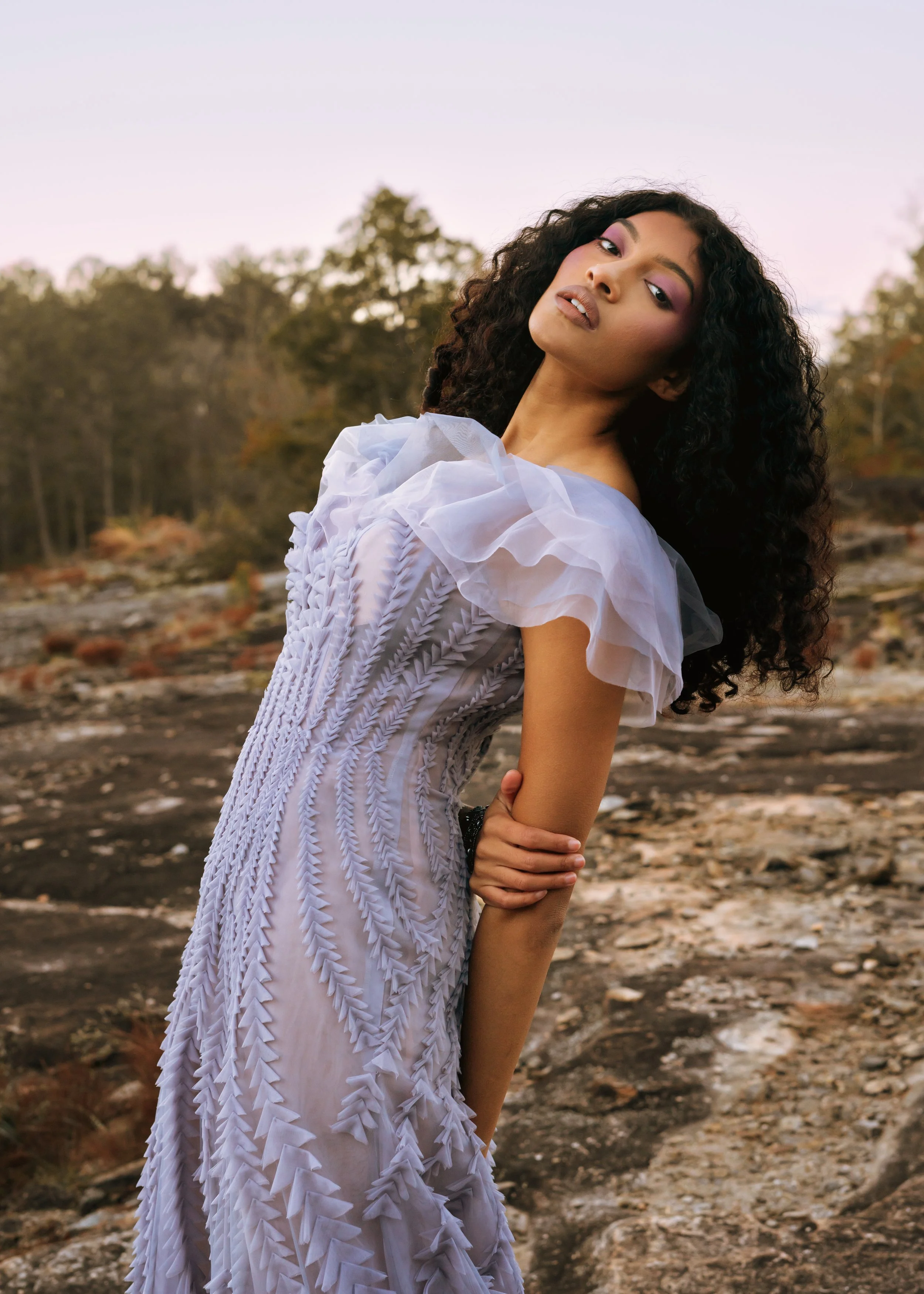 A woman with curly hair wearing a white dress with ruffled sleeves standing outdoors in a natural setting with trees and rocky ground during sunset.