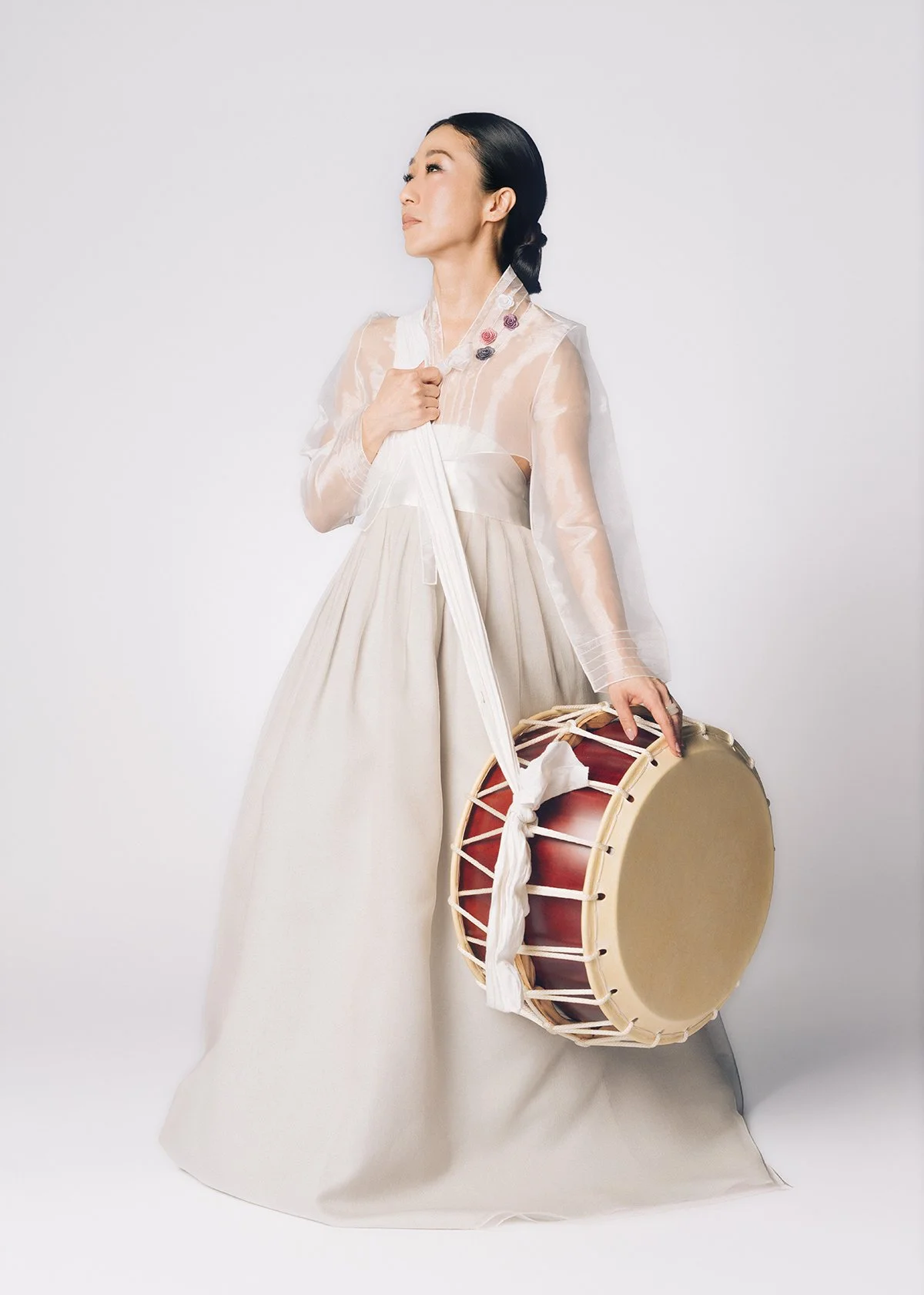 A woman dressed in traditional Korean hanbok holding a large drum with a drumstick.