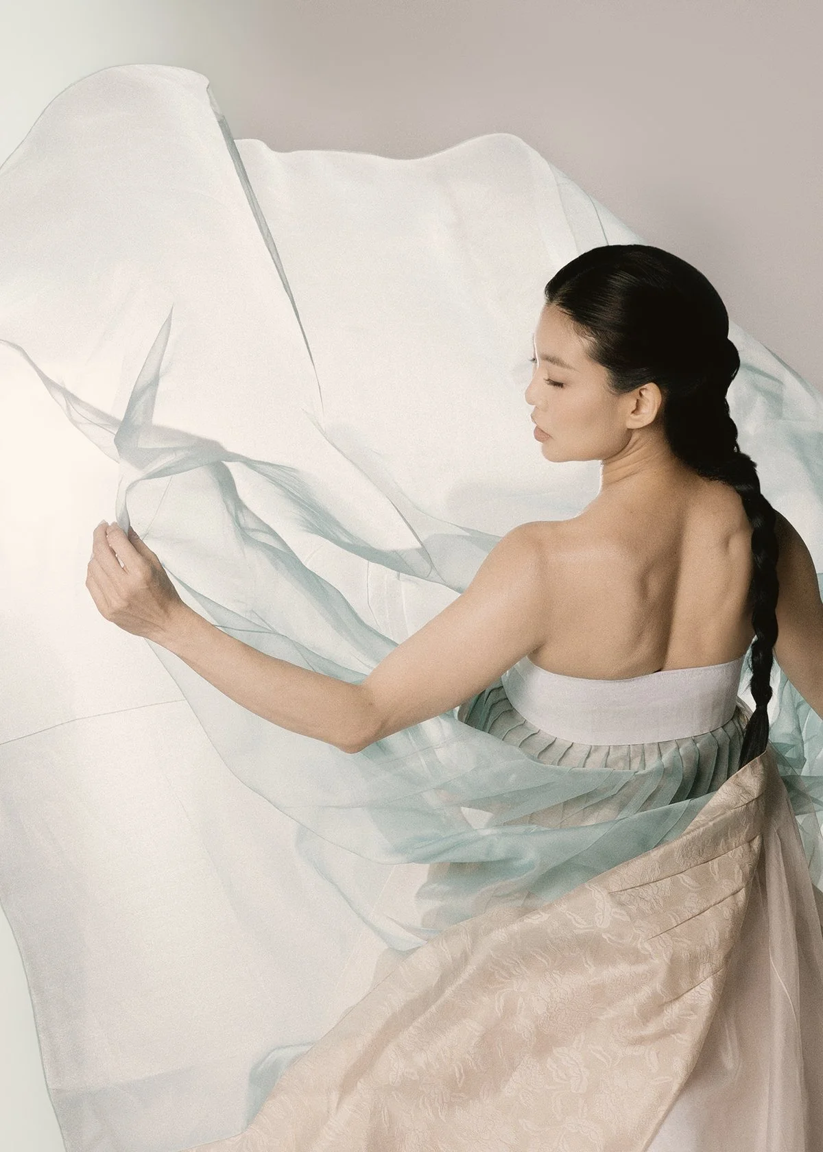 A woman with long dark hair in a braid, wearing a strapless gown, is sitting and holding a sheer fabric, creating flowing shapes around her.