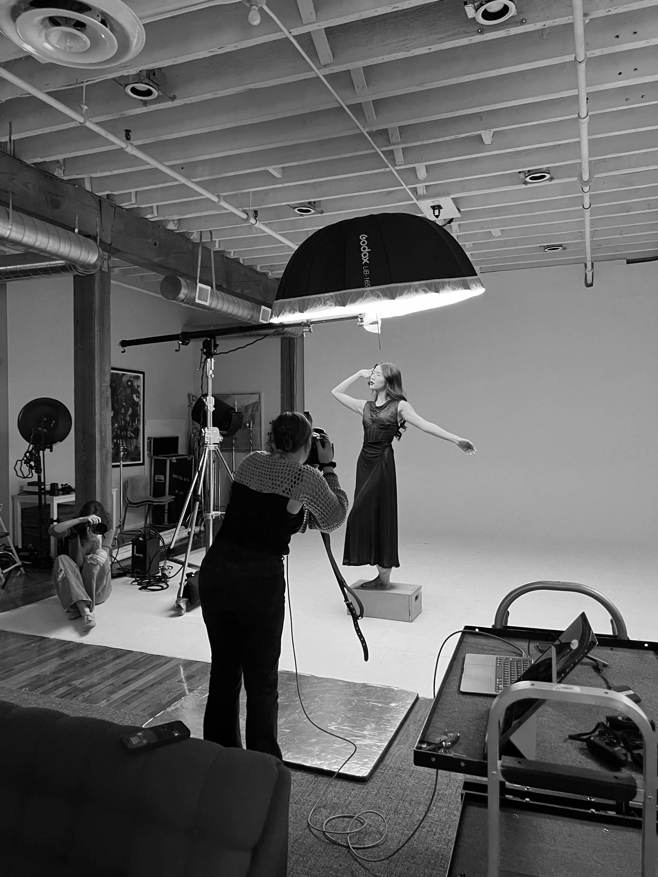 A professional photoshoot in a studio with a woman model in a dress posing on a platform. Photographers are taking photos while using studio lighting and equipment, including a large umbrella light overhead.