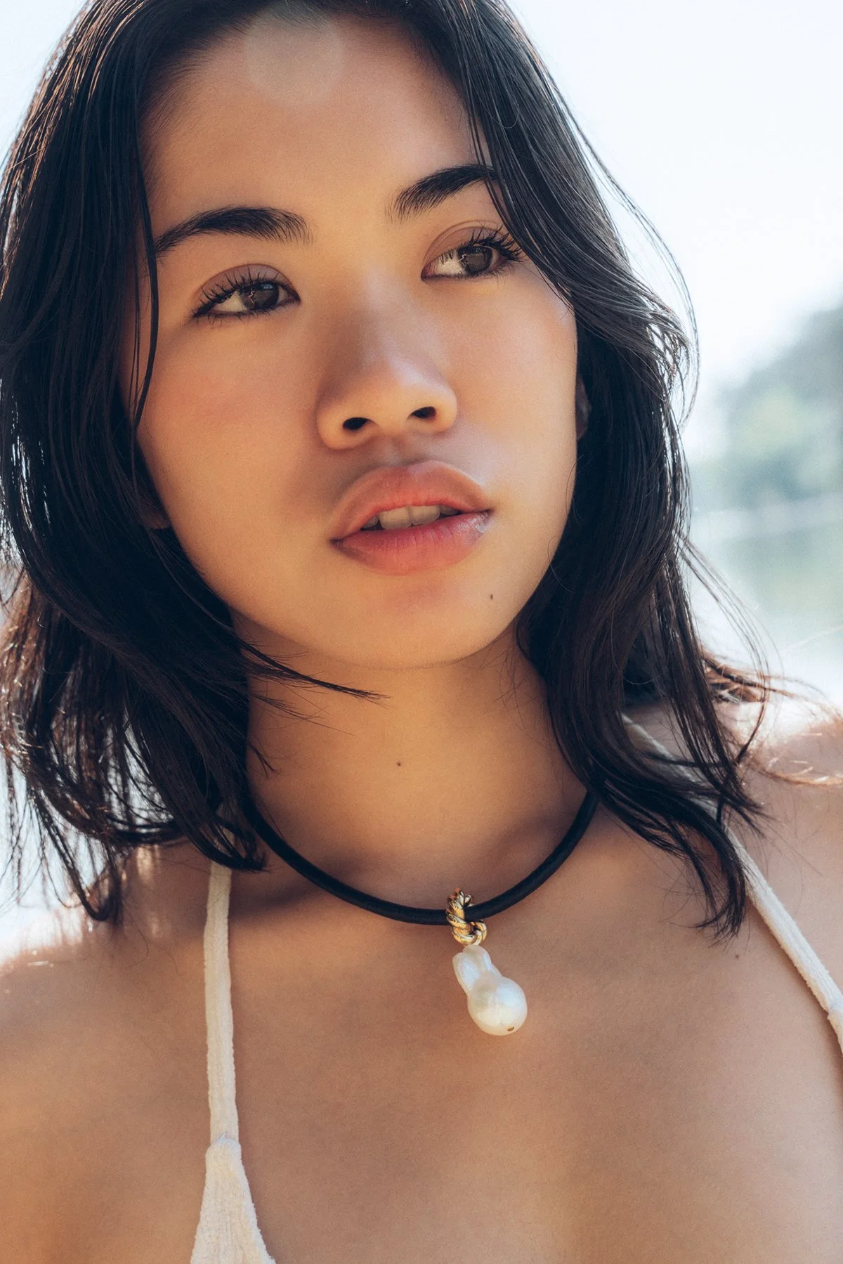 Close-up of a young woman with shoulder-length black hair, wearing a black choker with a pearl pendant, and a white strap top. The background is bright and out of focus.