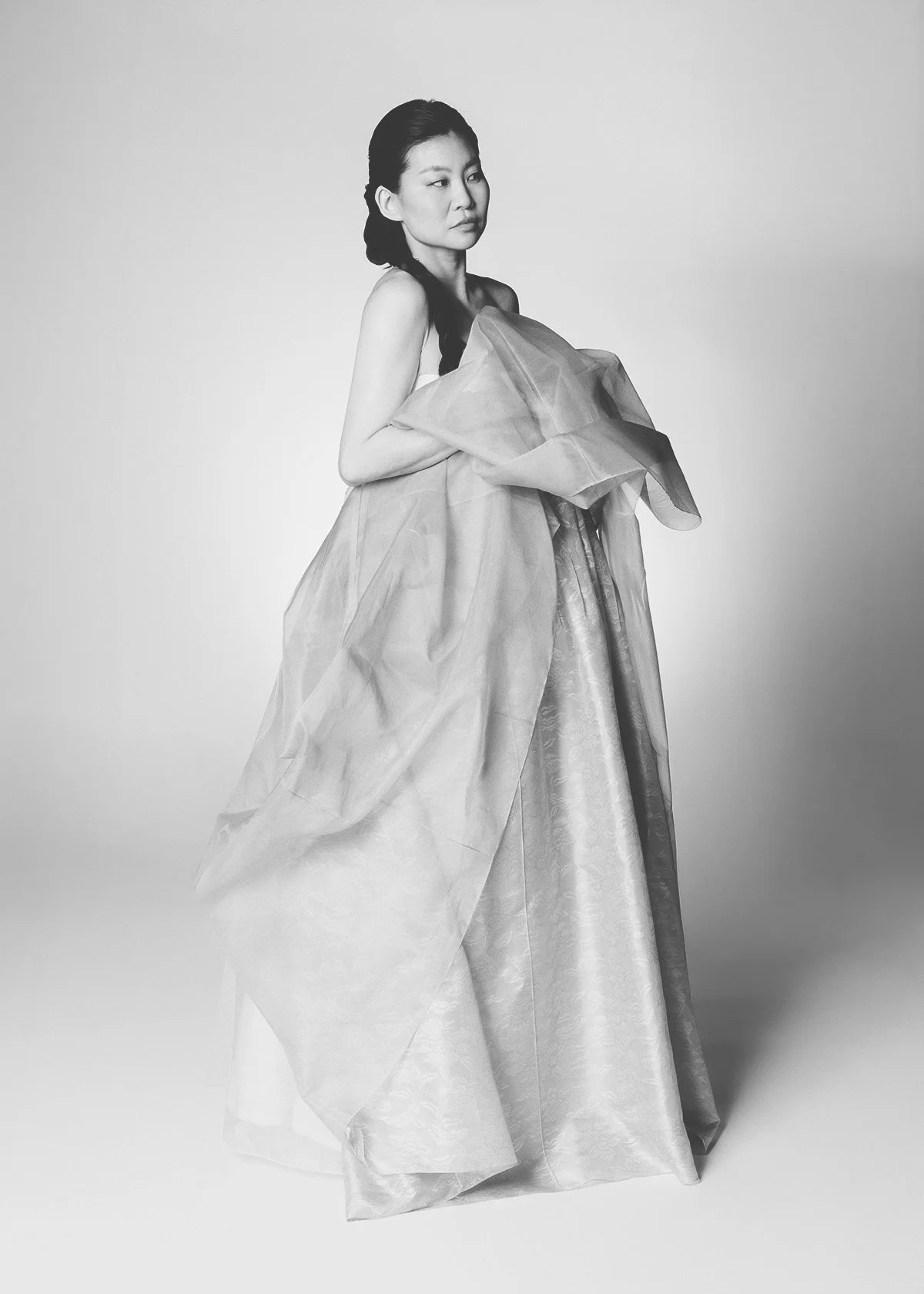 Black-and-white photo of a woman in an elegant, flowing gown holding fabric, looking to her right, against a plain background.