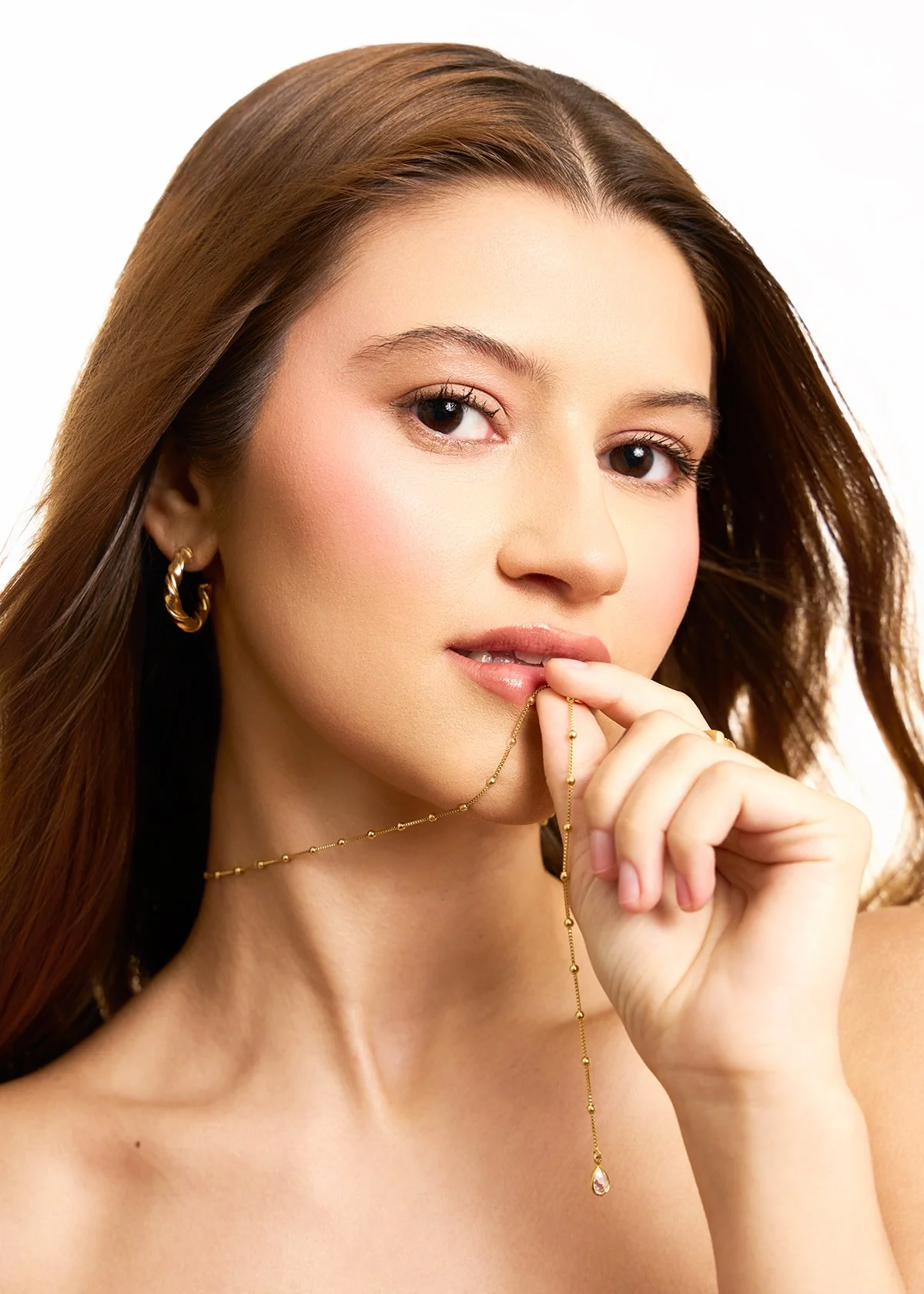 Close-up of a young woman with long brown hair, gold hoop earrings, and delicate gold jewelry, holding a gold chain with a pendant near her lips, looking at the camera with a soft expression.
