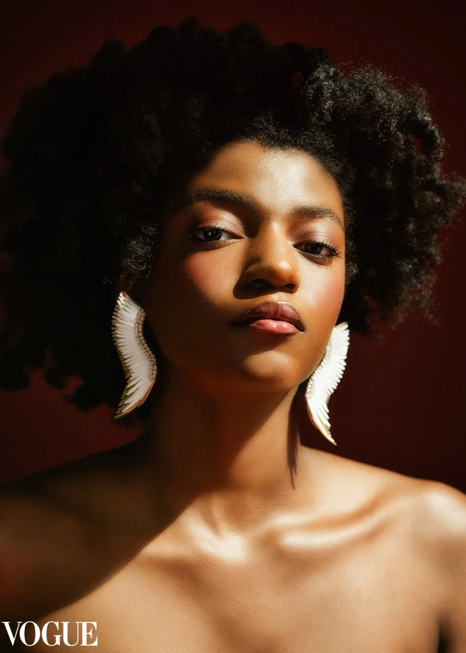 A close-up portrait of a woman with natural curly hair, wearing large white earrings, and a neutral expression, posing against a dark background, in a Vogue photoshoot.