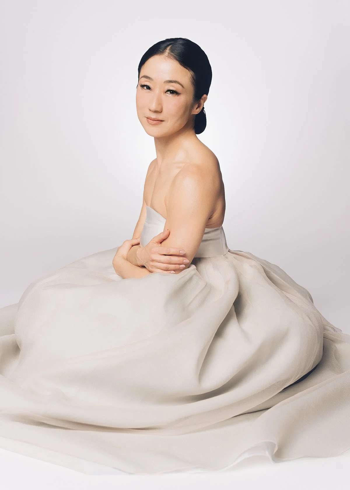A woman with dark hair styled in a sleek bun, sitting on the floor against a white background. She is wearing a strapless, light-colored, flowing dress and has her arms crossed over her knees, with a calm and gentle expression.
