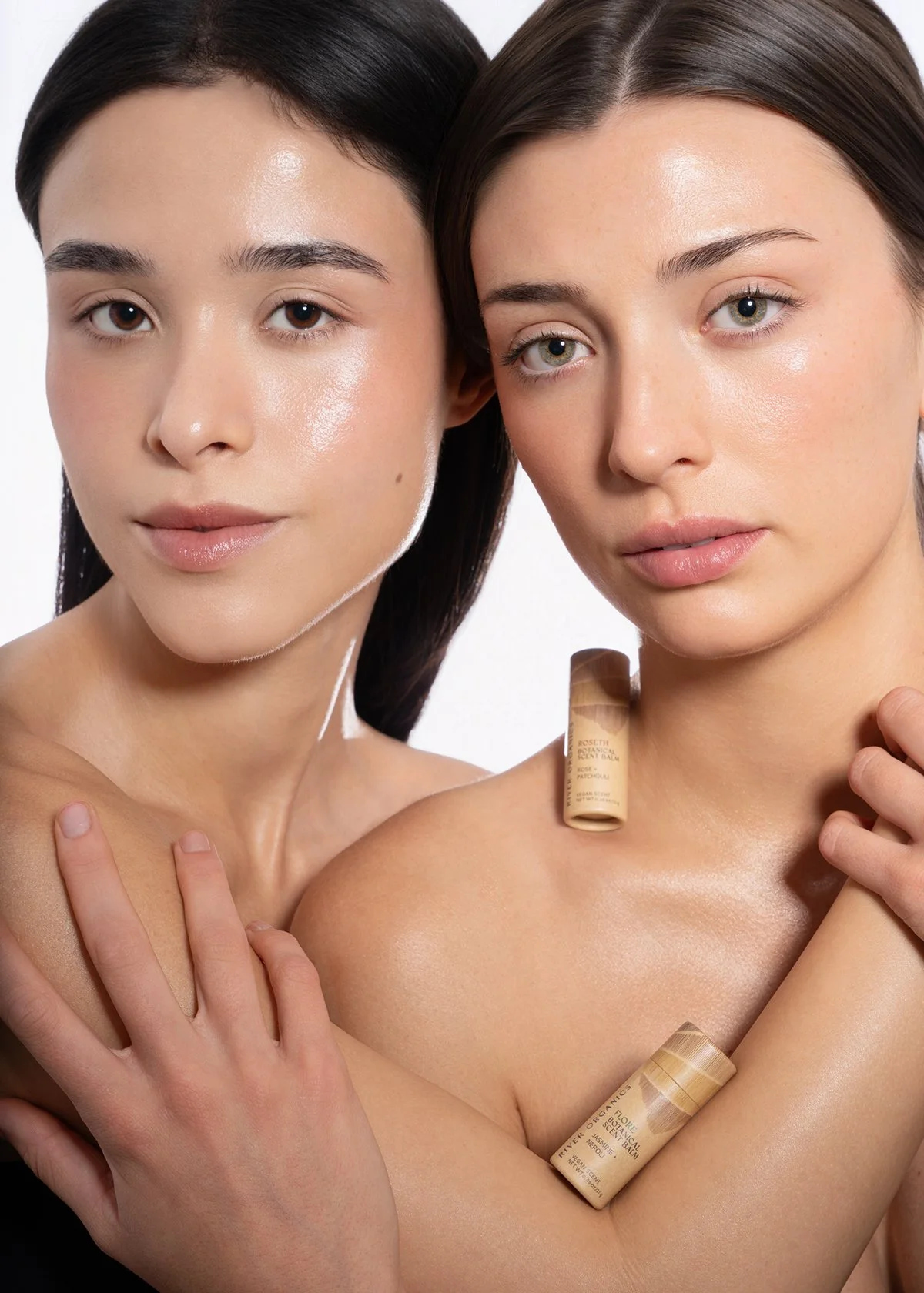 Close-up of two women with radiant skin holding skincare products, with one woman gently touching the other's shoulder, against a plain white background.