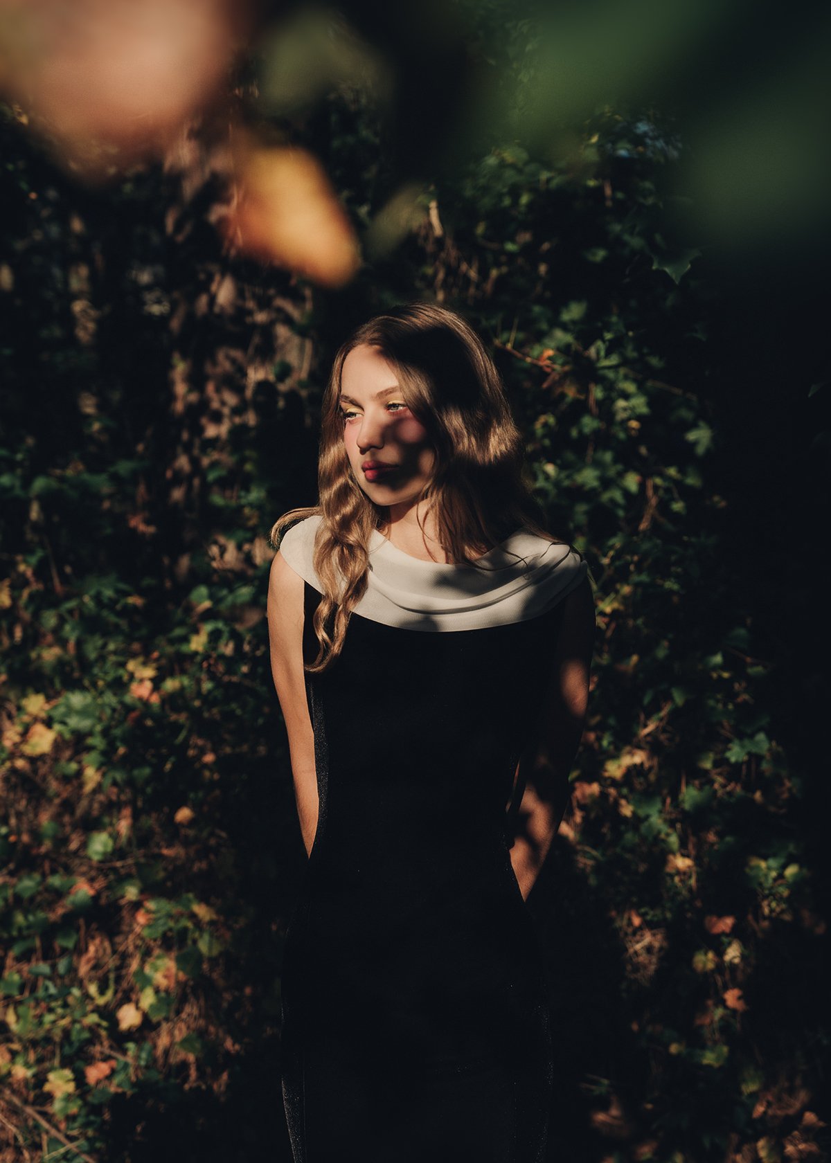 Young woman with wavy hair standing outdoors in a leafy area, partly shaded by branches.