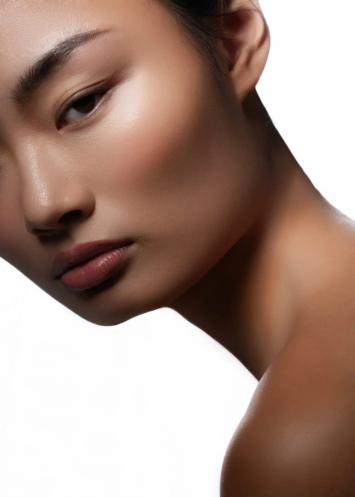 Close-up of a woman's face with smooth skin, subtle makeup, and a neutral expression, showing her eye, cheek, lips, and shoulder against a white background.