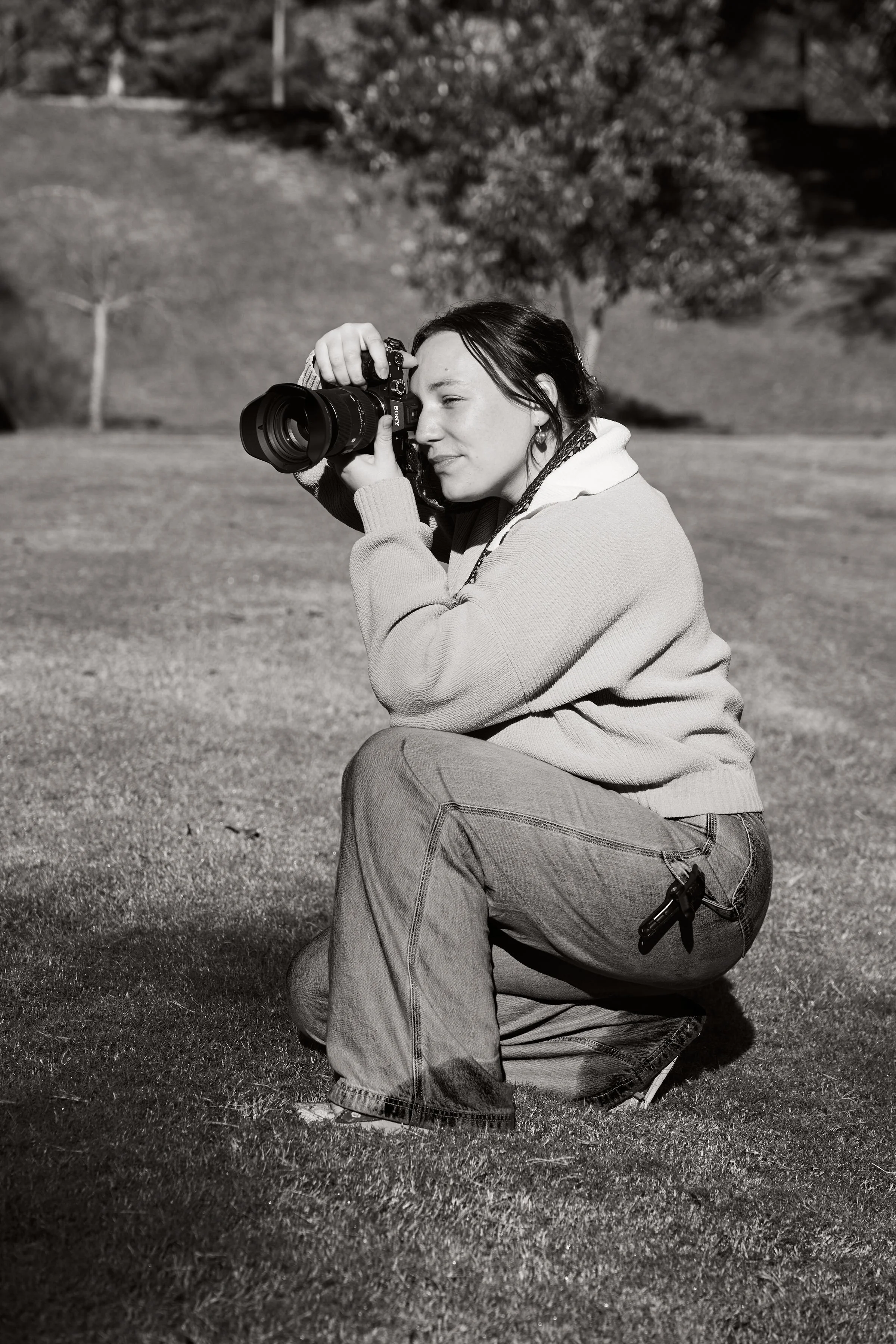 A woman crouching on the grass, taking a photograph with a DSLR camera, outdoors in a park or garden setting.