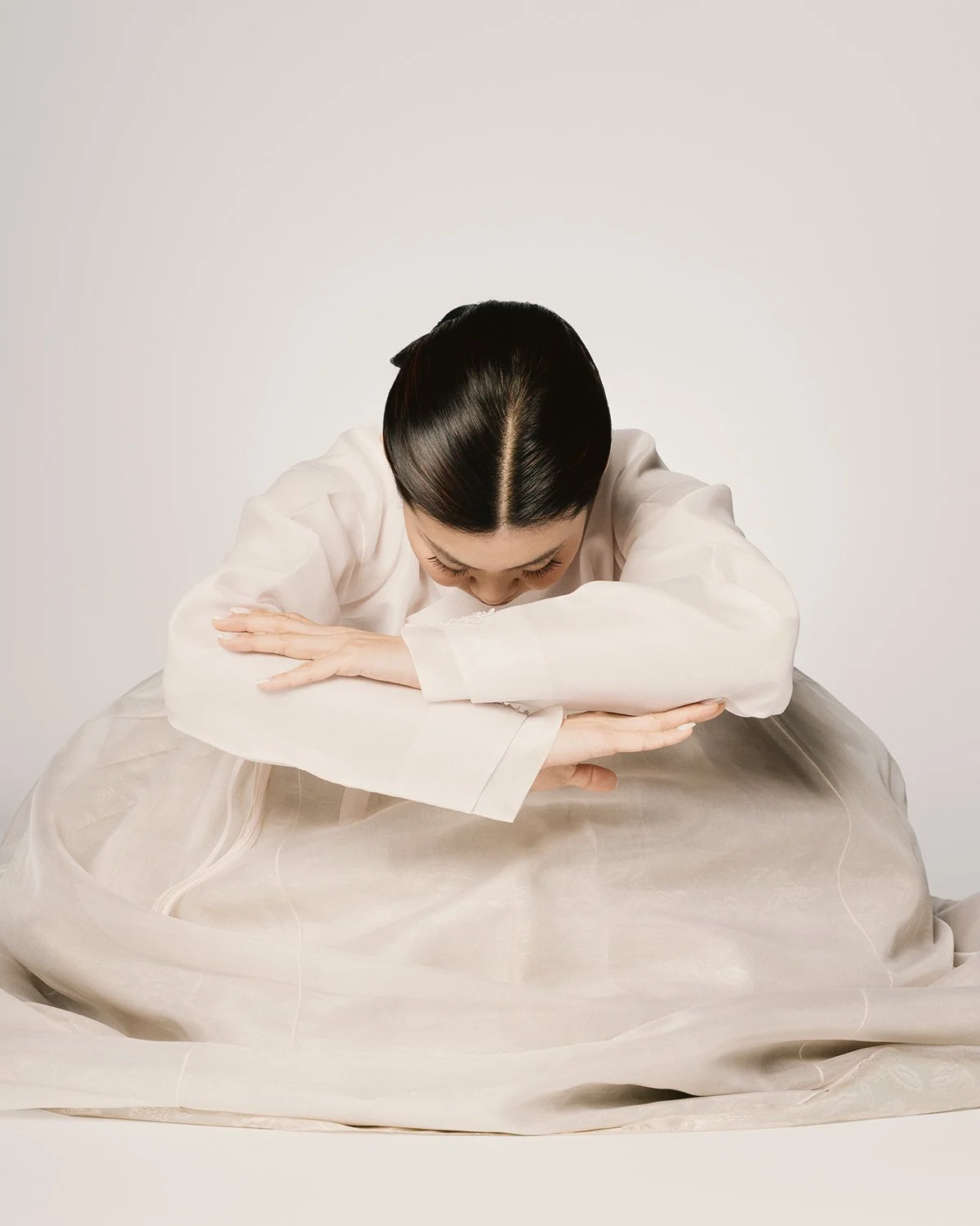 Woman with dark hair styled in a sleek bun, bowing her head and clasping her arms while wearing a white long-sleeve blouse and sitting on a large beige cushion or fabric in a neutral setting.